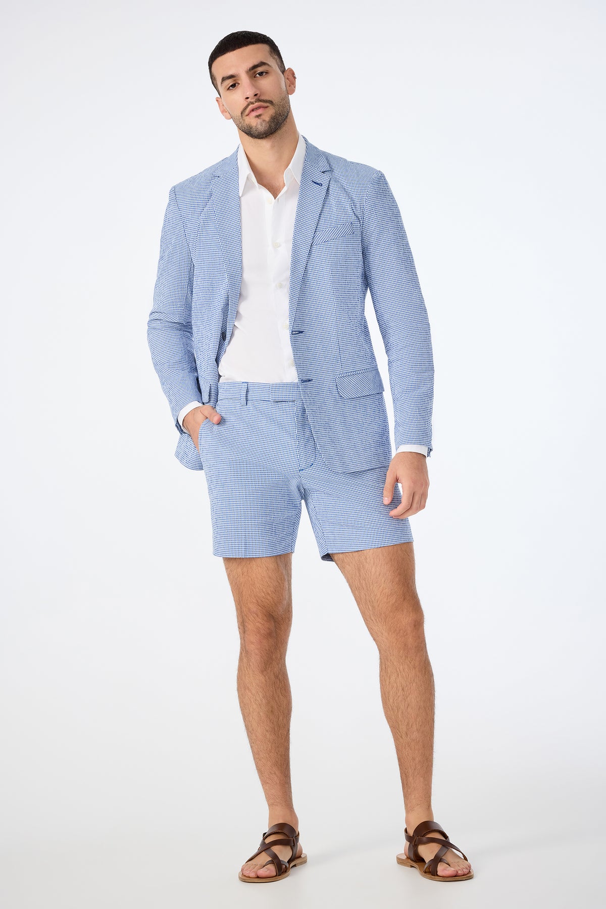 A man poses confidently in the Roger Short - Ocean Gingham set: a light blue seersucker suit with matching shorts, paired with a white dress shirt and brown sandals, against a plain white background.