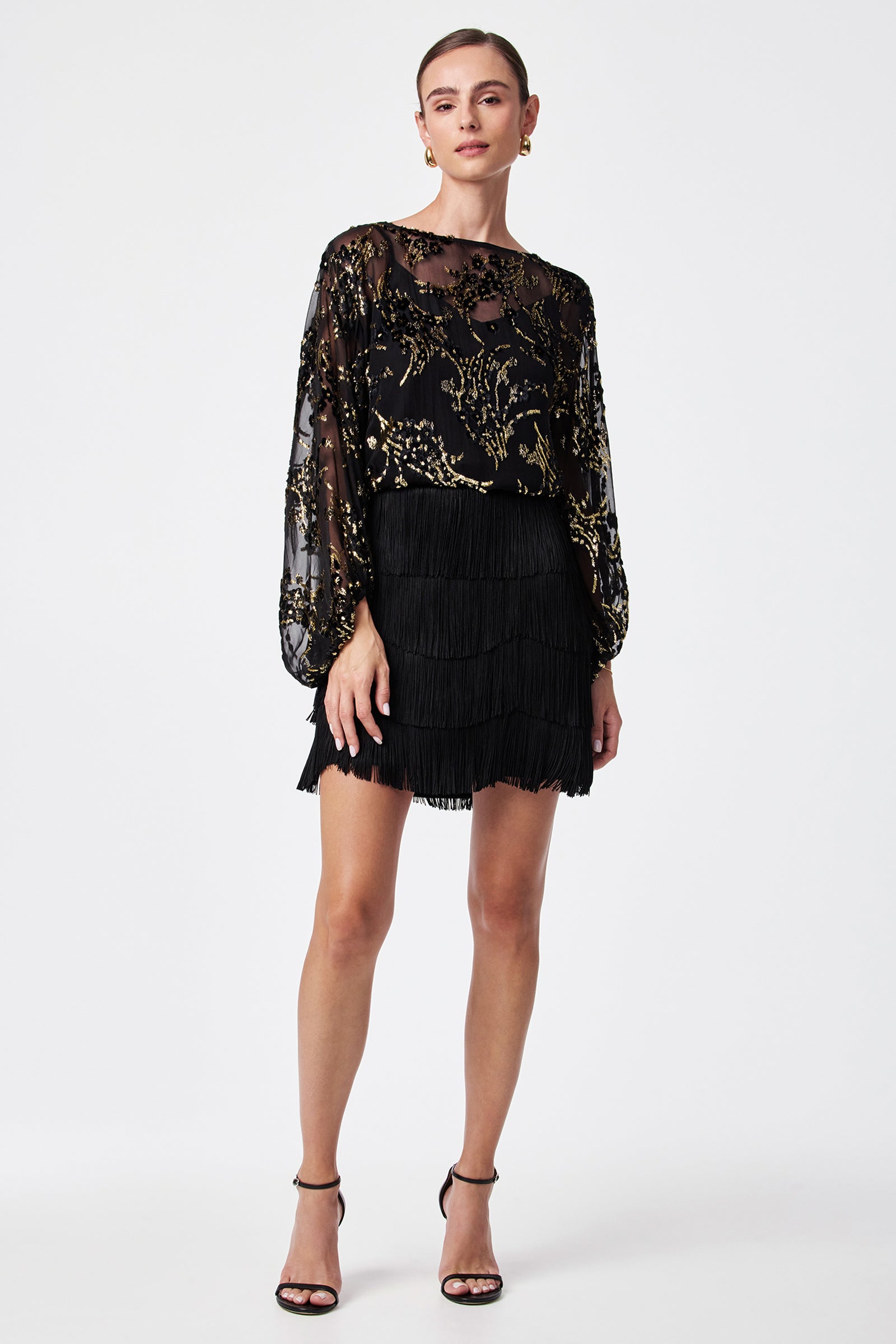 A woman stands against a white background wearing the Ursa Metallic Burnout Balloon Sleeve Top, a black tiered fringe skirt, black high-heeled sandals, and gold hoop earrings. Her hair is pulled back.