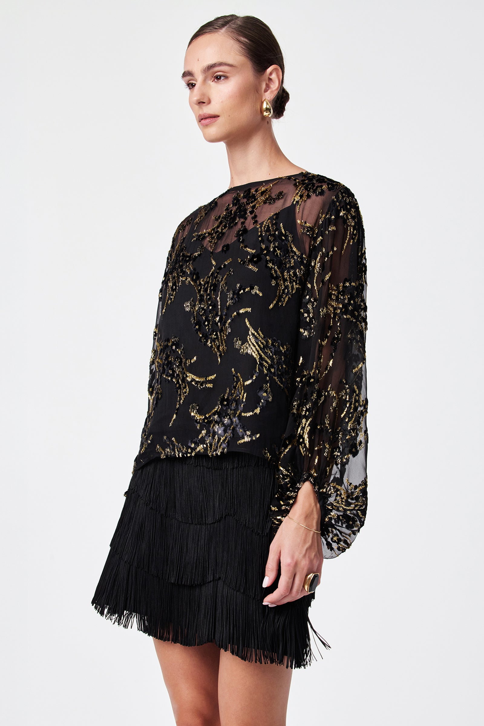 Wearing the Ursa Metallic Burnout Balloon Sleeve Top—a sheer black top with gold sequin patterns—paired with a black fringed mini skirt, gold hoop earrings, and a gold ring, against a plain white background.