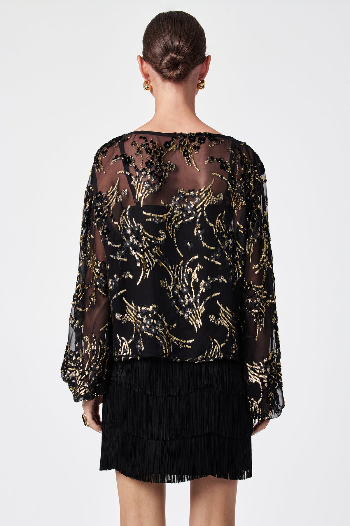 A woman with her hair in a bun is shown from the back, wearing the Ursa Metallic Burnout Balloon Sleeve Top and a short black fringed skirt, accessorized with gold hoop earrings.