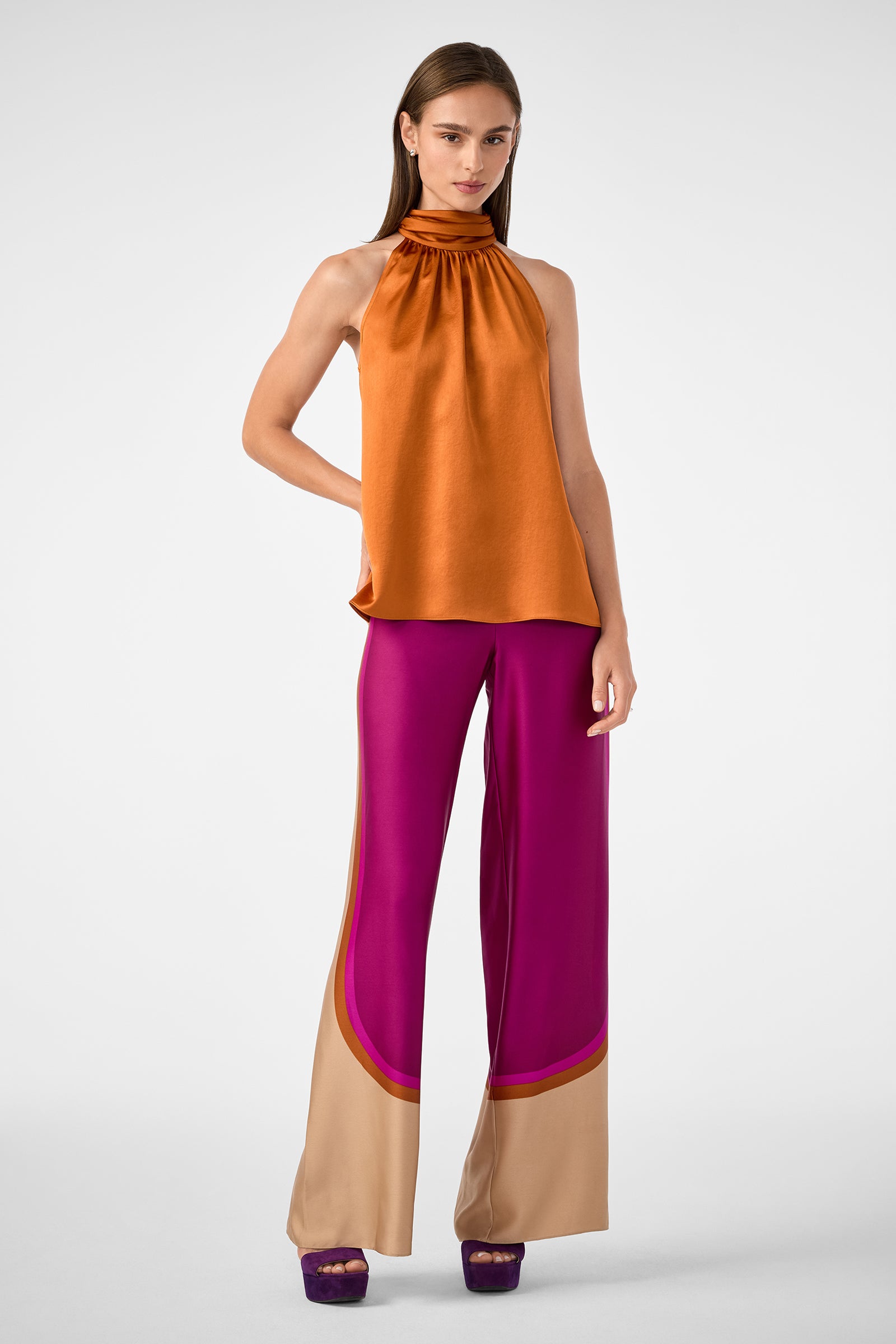 A woman wears the Verity 2 Pant—wide-legged magenta pants with beige and magenta hem accents—styled with an orange halter-neck top and purple platform heels, posing against a plain light background.