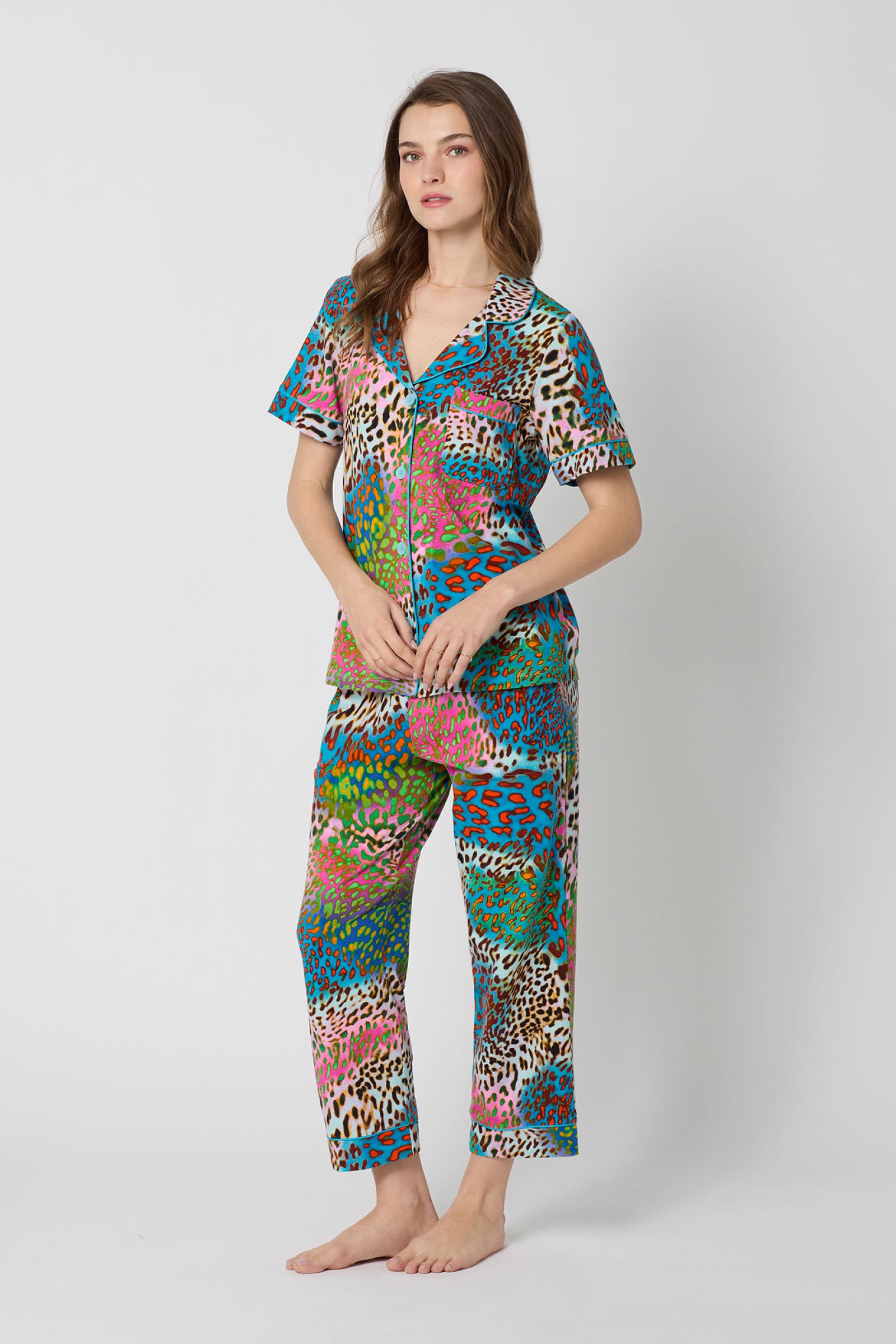 A woman stands barefoot against a plain background wearing the Crop Classic PJ Set - Spray Leopard, a colorful short-sleeved pajama set featuring a bold multicolored animal print with blue, pink, green, and brown cheetah spots.