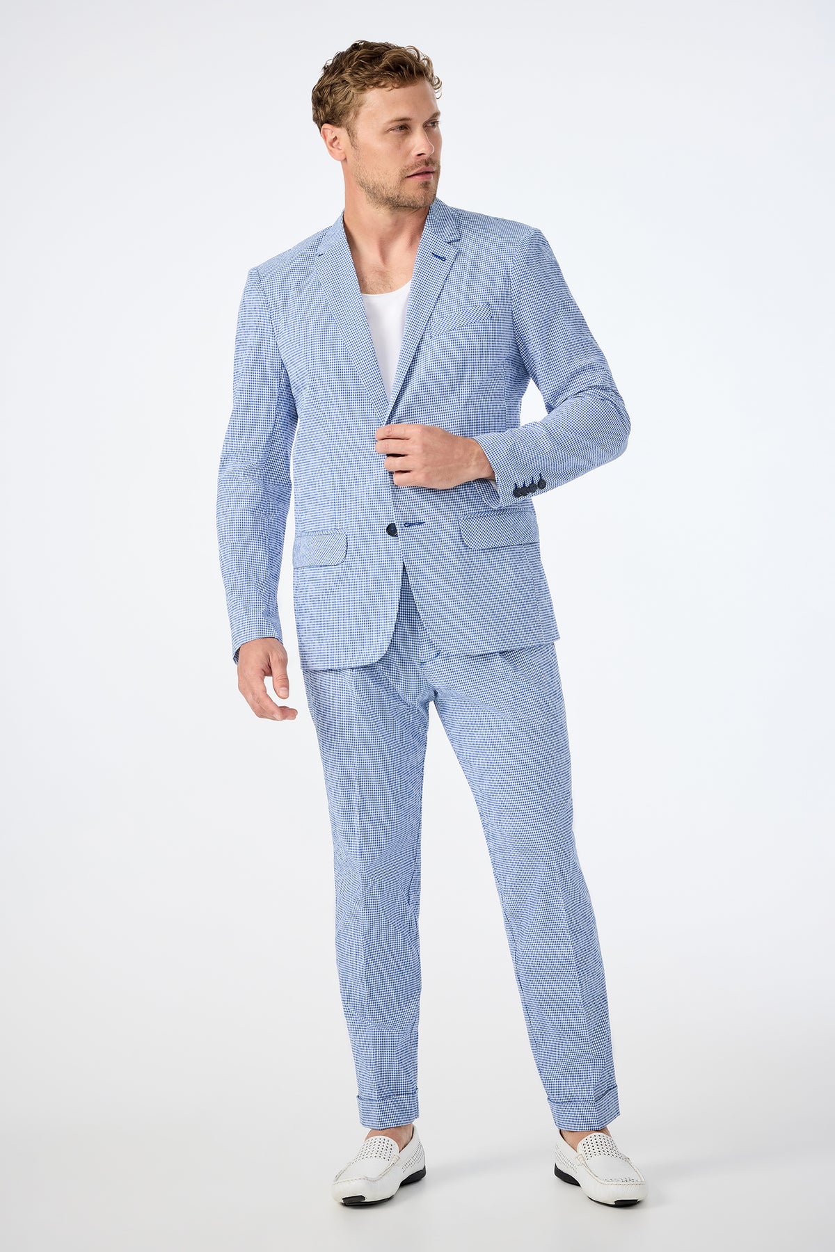 A man wears the Beale Pleated Trouser in Ocean Gingham with a matching jacket, white T-shirt, and white loafers, standing against a plain white background and looking to the side.