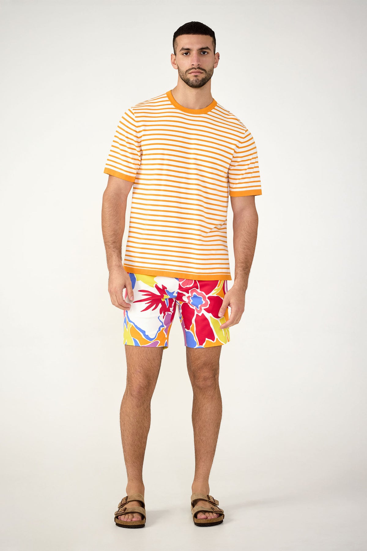 A man with short dark hair and a trimmed beard wears brown sandals, a yellow and white striped t-shirt, and Lawrence Short - Soi Paradis: colorful floral shorts in red, yellow, blue, and white against a plain background.