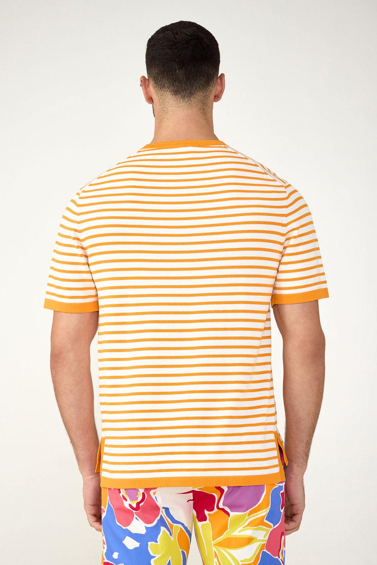 A man is seen from behind wearing the Giovanni Stripe Short-Sleeve Pullover in orange and white, paired with vibrant floral shorts featuring red, blue, white, and yellow patterns against a plain light background.