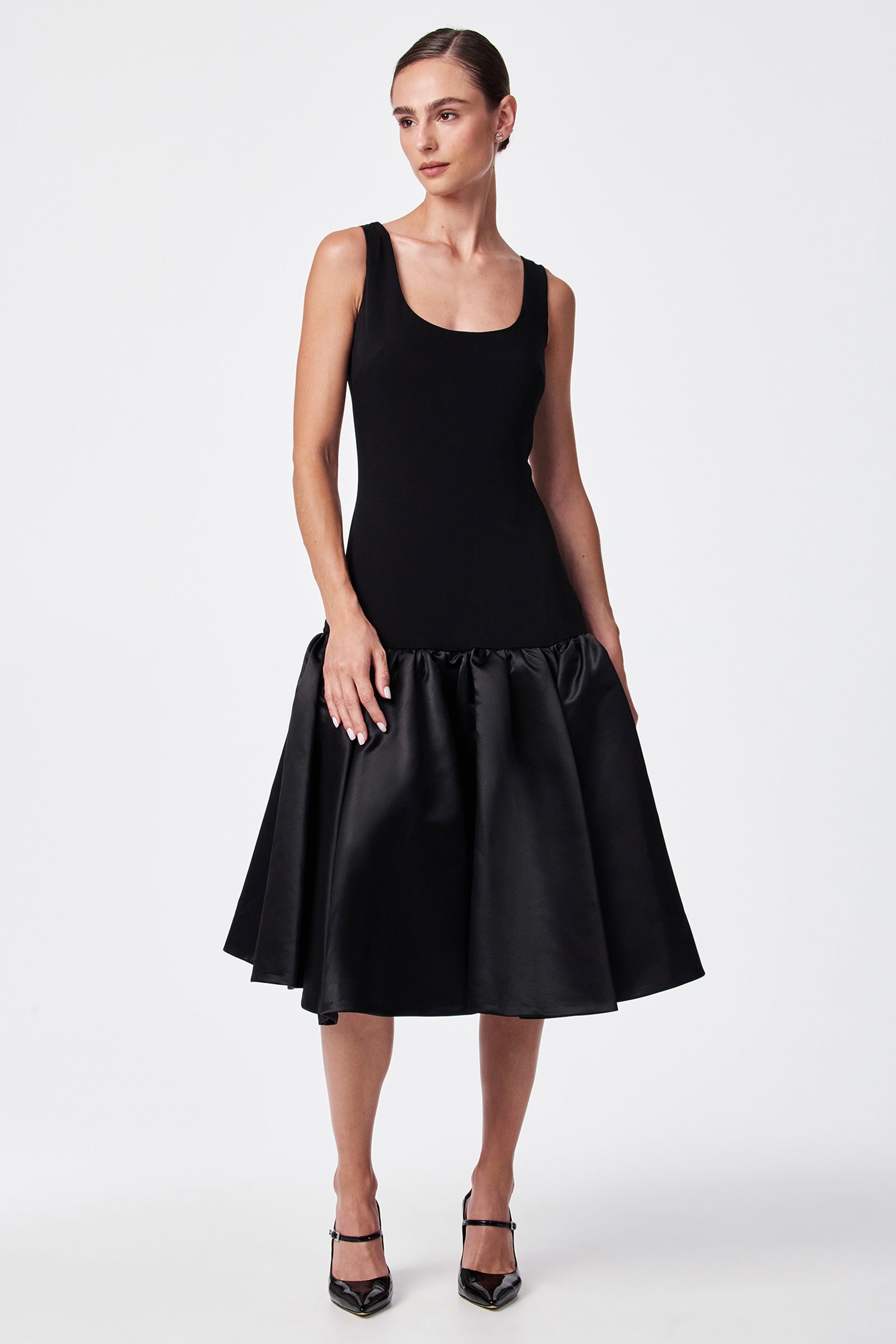 A woman stands against a plain white background wearing the SINA DRESS - BLACK, a sleeveless dress with a fitted bodice and flared satin skirt, paired with black pointed-toe heels.