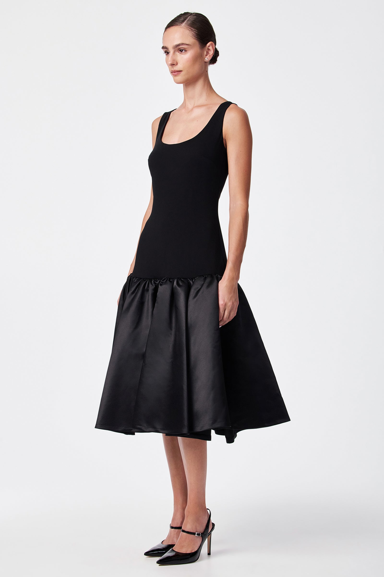 A woman with her hair in a sleek bun wears the SINA DRESS - BLACK, featuring a sleeveless fitted top and flared shin-length satin skirt, paired with black high heels, standing against a plain white background.