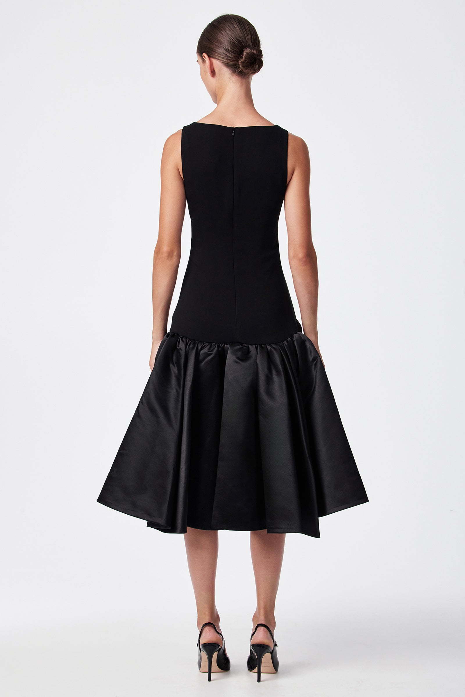 A woman stands facing away in the SINA DRESS - BLACK, a sleeveless dress with a fitted top and ruffled, voluminous skirt. Her hair is in a neat bun and she wears black high-heeled sandals against a white background.