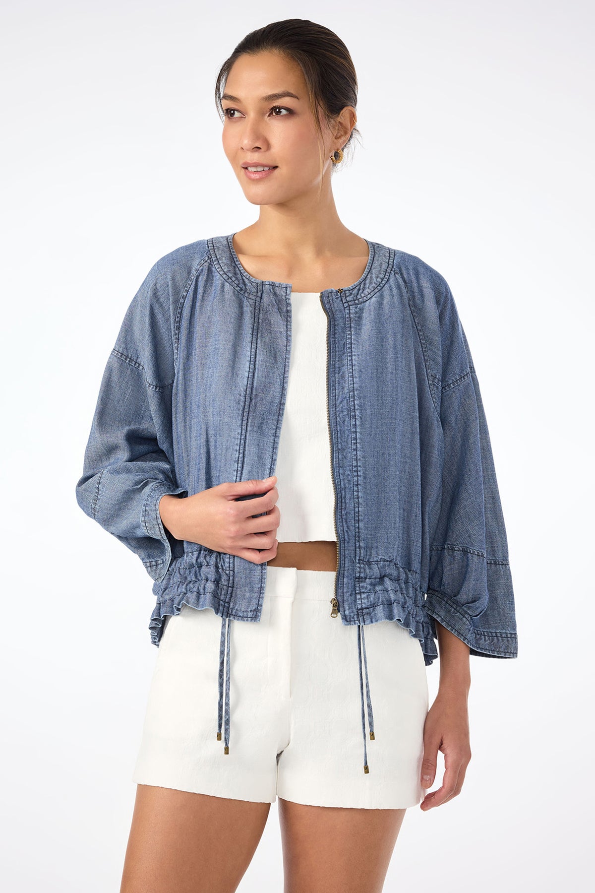 A woman stands against a plain background wearing the Prita Chambray Jacket in indigo over a white top and white shorts, looking slightly to the side and smiling softly.