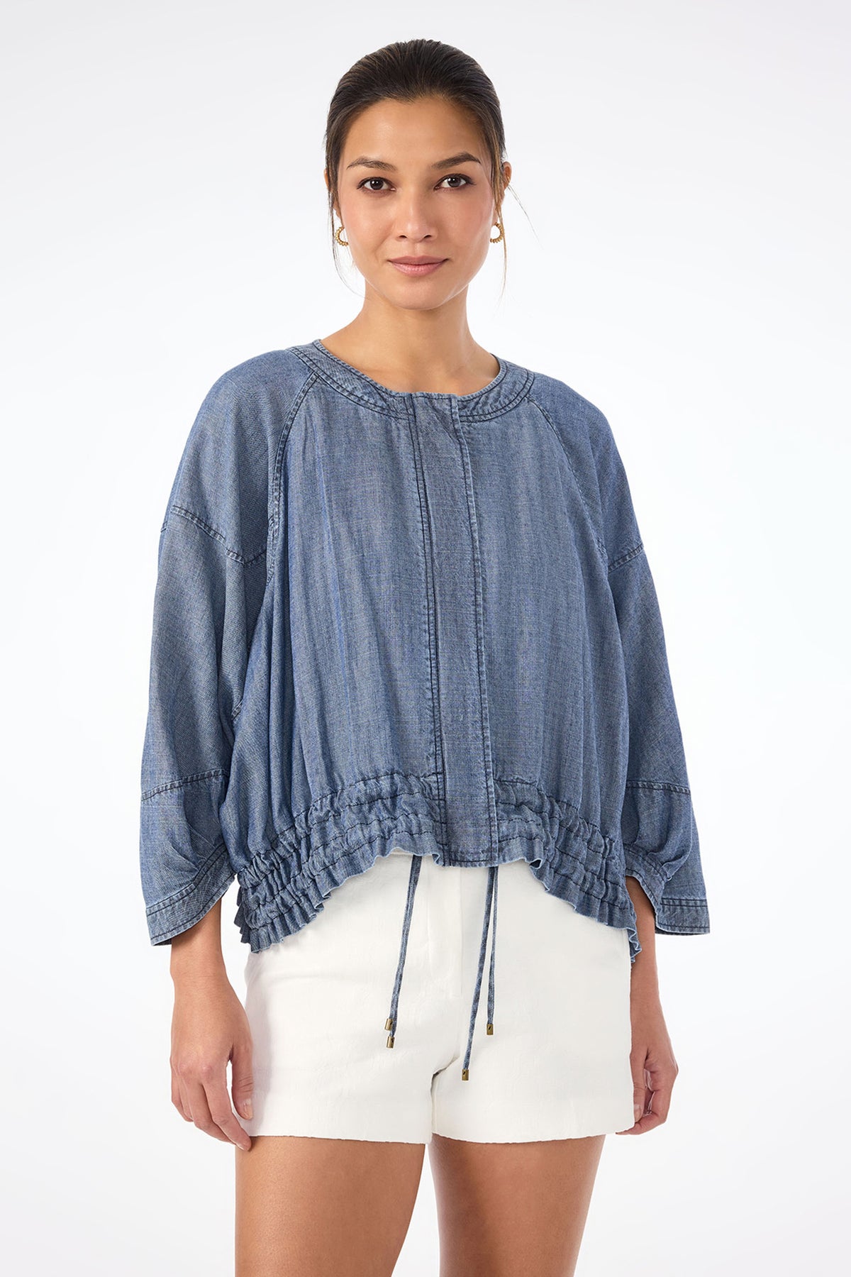 A woman faces forward, wearing the Prita Chambray Jacket‚Äö√Ñ√Æa loose-fitting blue denim blouse with gathered drawstrings at the hem‚Äö√Ñ√Æpaired with white shorts against a plain white background.