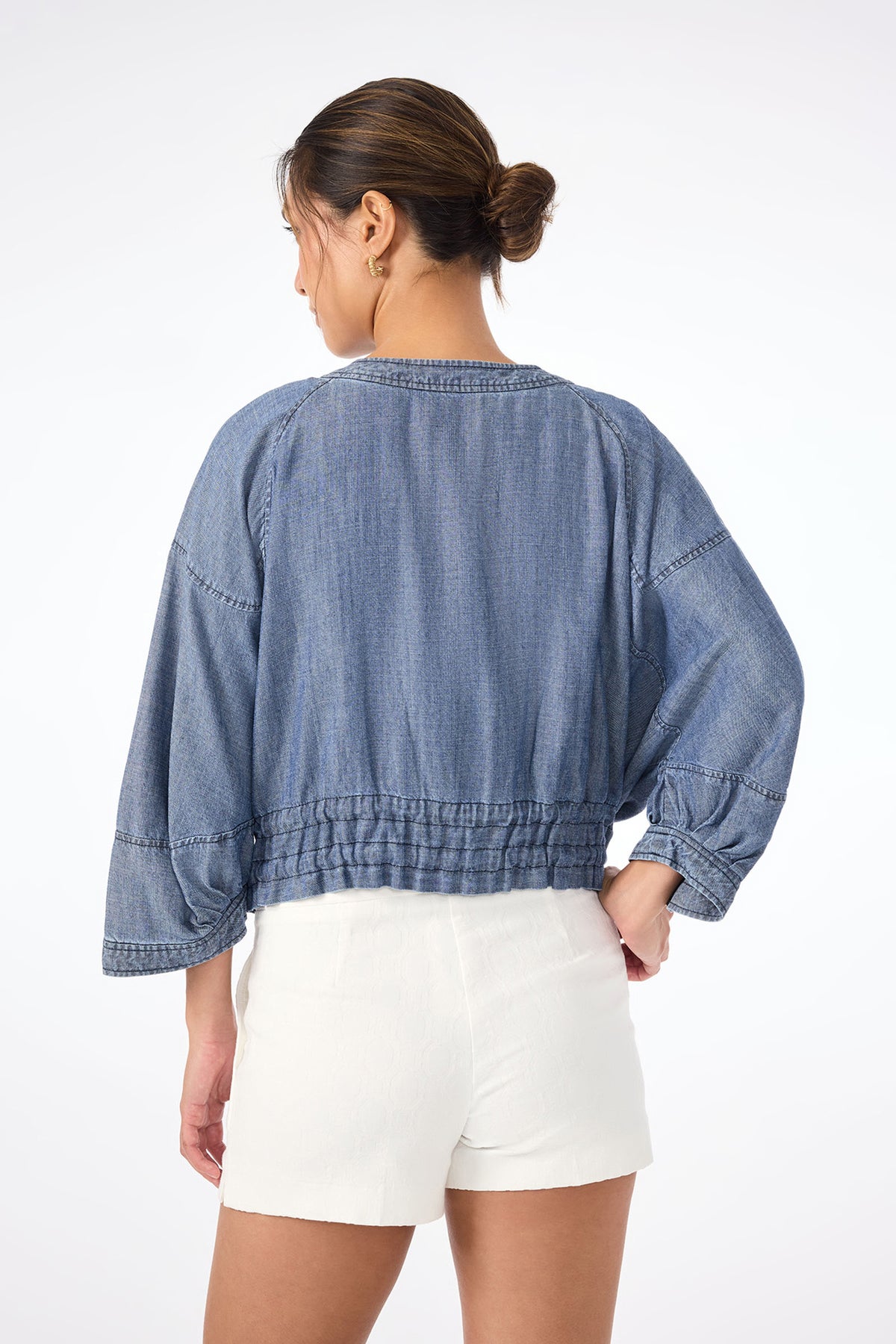 A woman with her hair in a neat bun faces away, wearing the loose-fitting Prita Chambray Jacket with gathered hem and white high-waisted shorts against a plain white background.