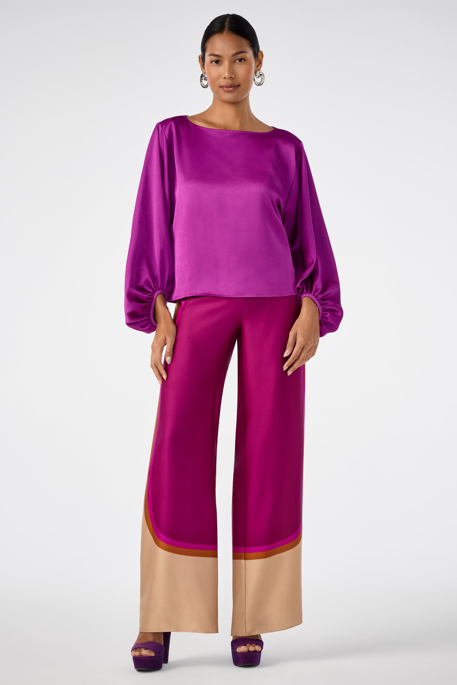 A woman stands wearing the shiny magenta Ursa 2 Top with matching wide-leg magenta pants featuring tan and red trim at the hem and purple heels, set against a plain light background.