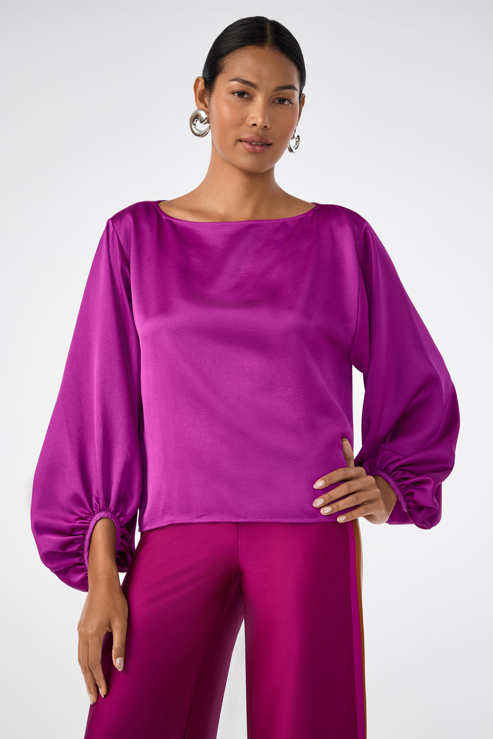 A woman stands confidently in the Ursa 2 Top, a shiny magenta long-sleeve blouse with voluminous sleeves, paired with matching pants. She accessorizes with silver hoop earrings and has her dark hair pulled back against a light gray background.