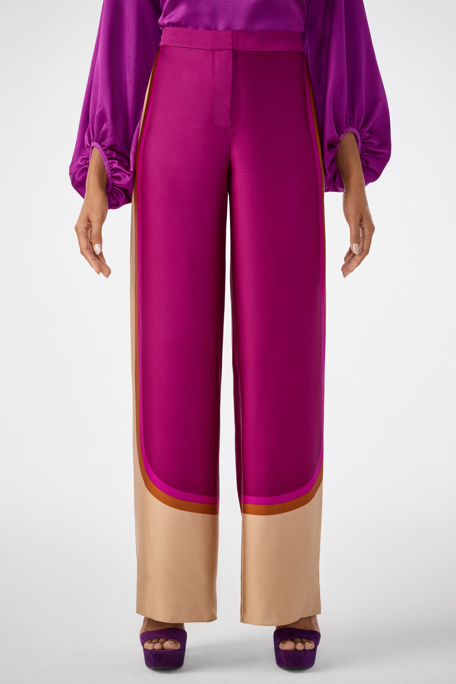 A person wears the Verity 2 Pant in magenta with tan and brown trim at the hem, paired with a matching magenta top featuring billowy sleeves and purple platform heels, set against a white background.