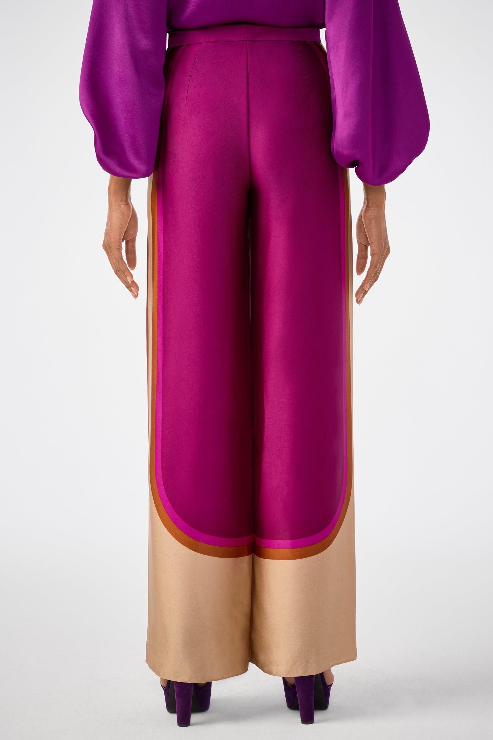 A person stands with their back to the camera, wearing the Verity 2 Pant—high-waisted, wide-leg magenta pants featuring tan borders and rounded magenta and gold-edged cuffs; their top has voluminous magenta sleeves.