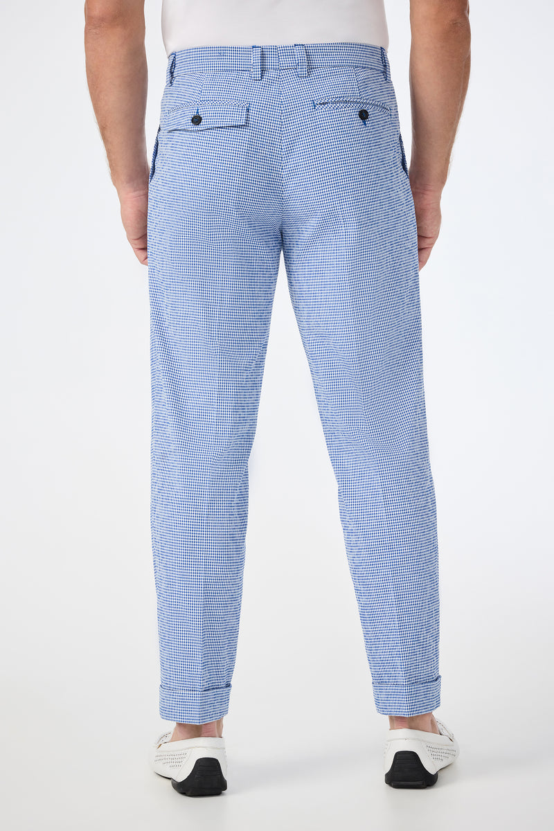 A man faces away, wearing Beale Pleated Trouser - Ocean Gingham: light blue small-check trousers with cuffed hems, two back pockets with dark buttons, paired with a white belt and sneakers. The background is plain white.