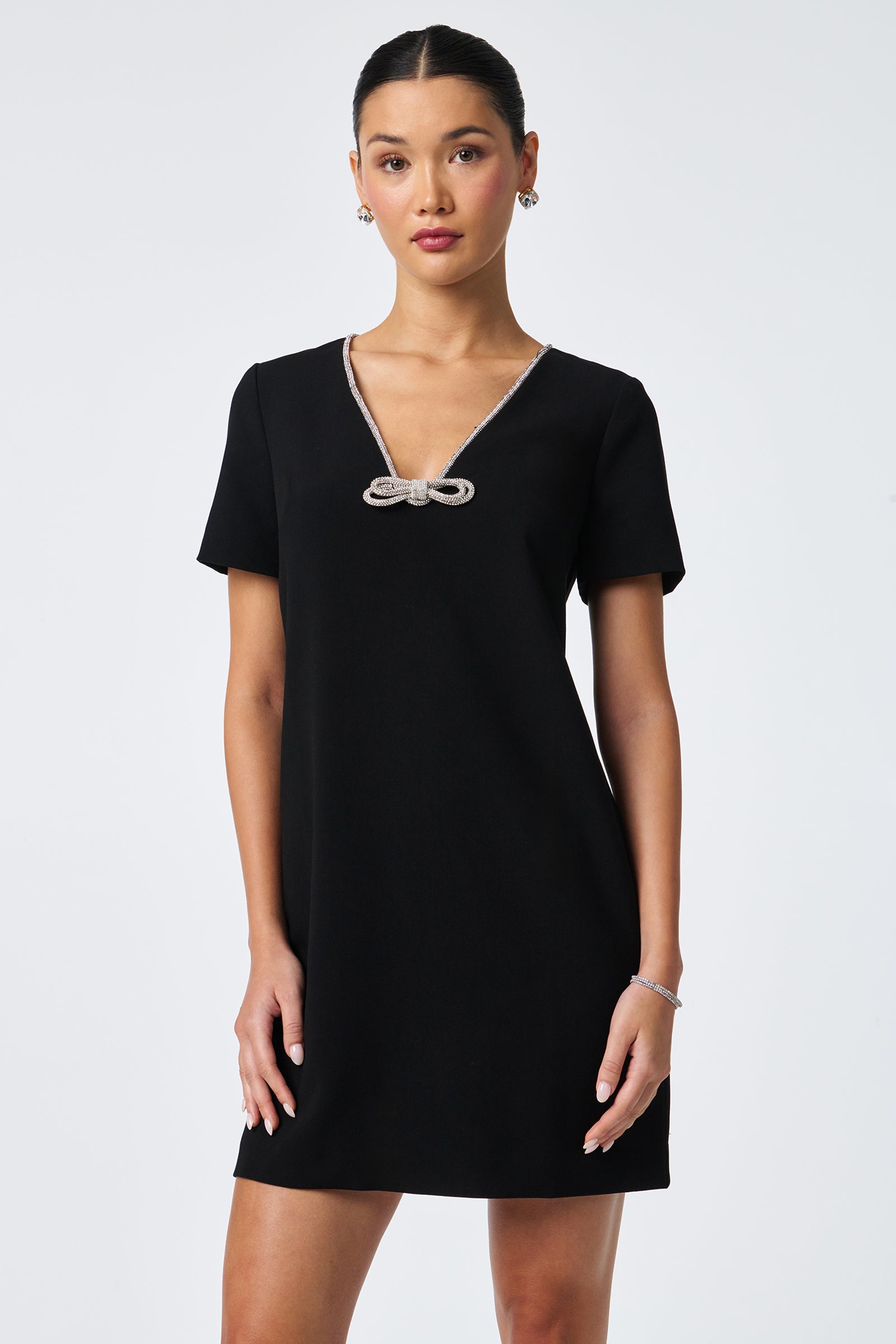 A woman stands against a white background wearing the Obelle Crystal Trim Mini Dress—a short-sleeved black V-neck dress with a small bow detail. She has her hair tied back and accessorizes with subtle jewelry.