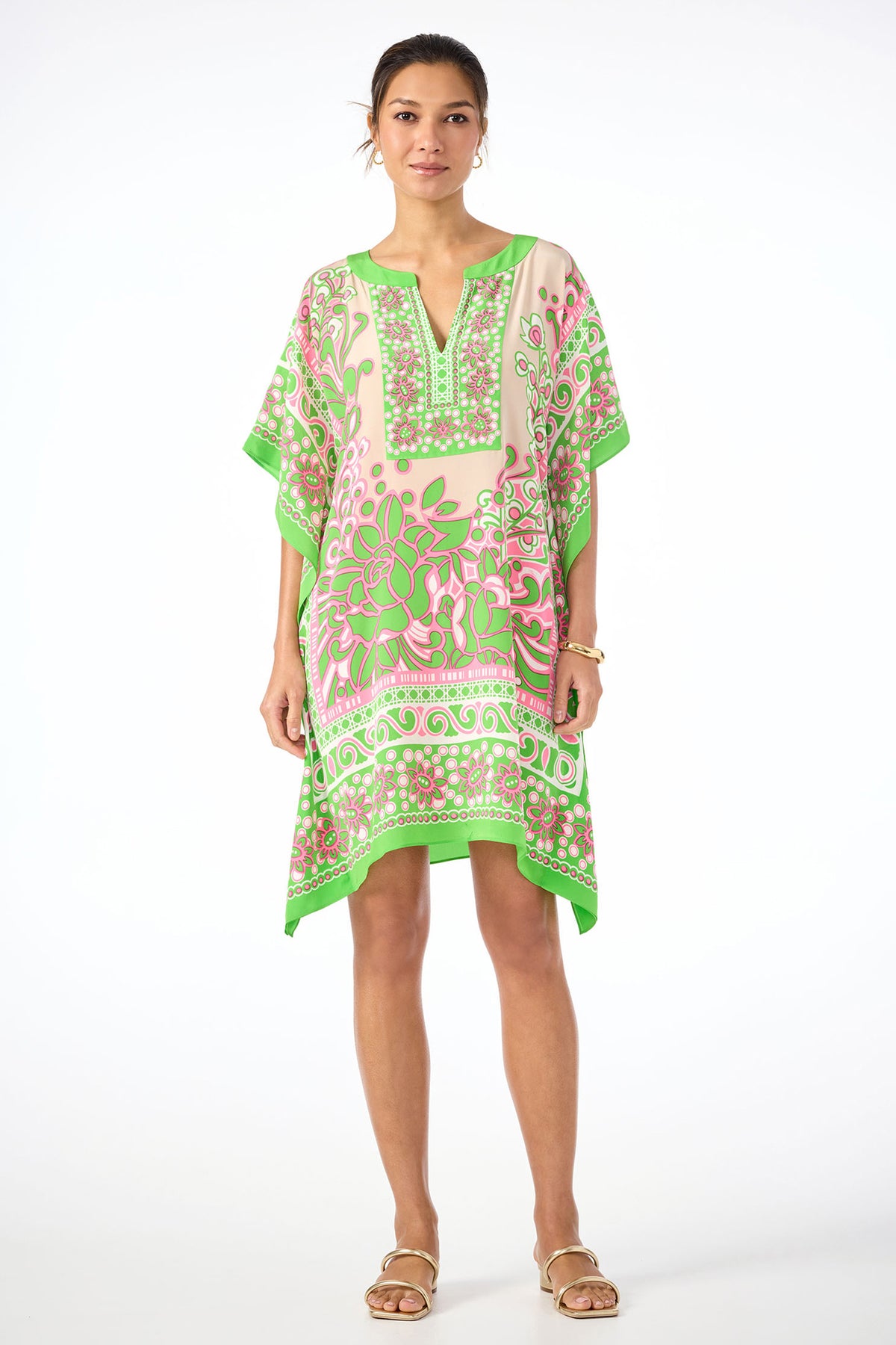 A woman wears the Theodora Caftan Dress in Petra Floral silk, a loose-fitting, knee-length dress with pink and green patterns, styled with bracelets and sandals against a white background.