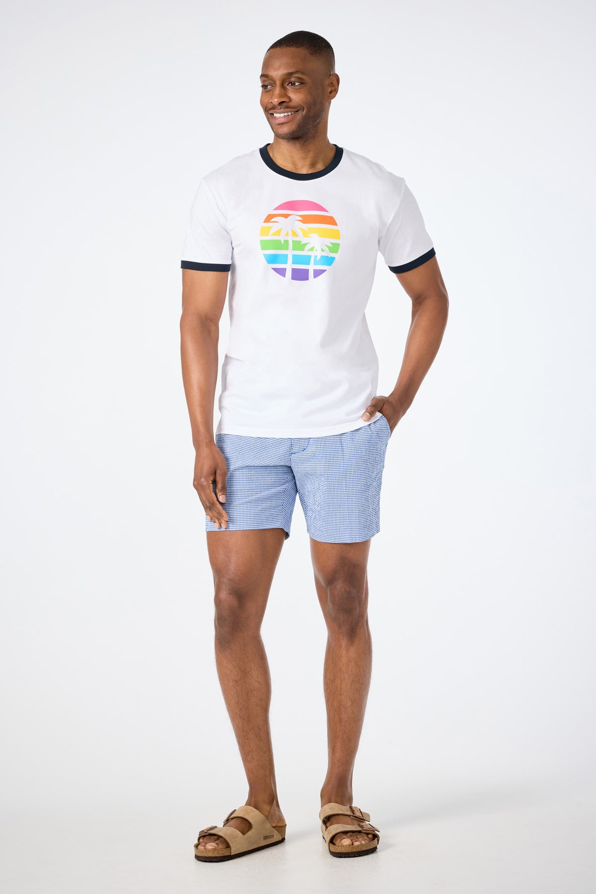 A man smiles in a white Short-Sleeve Crewneck T-Shirt - Pride featuring a rainbow palm tree graphic, paired with light blue shorts and brown sandals, against a plain white background.