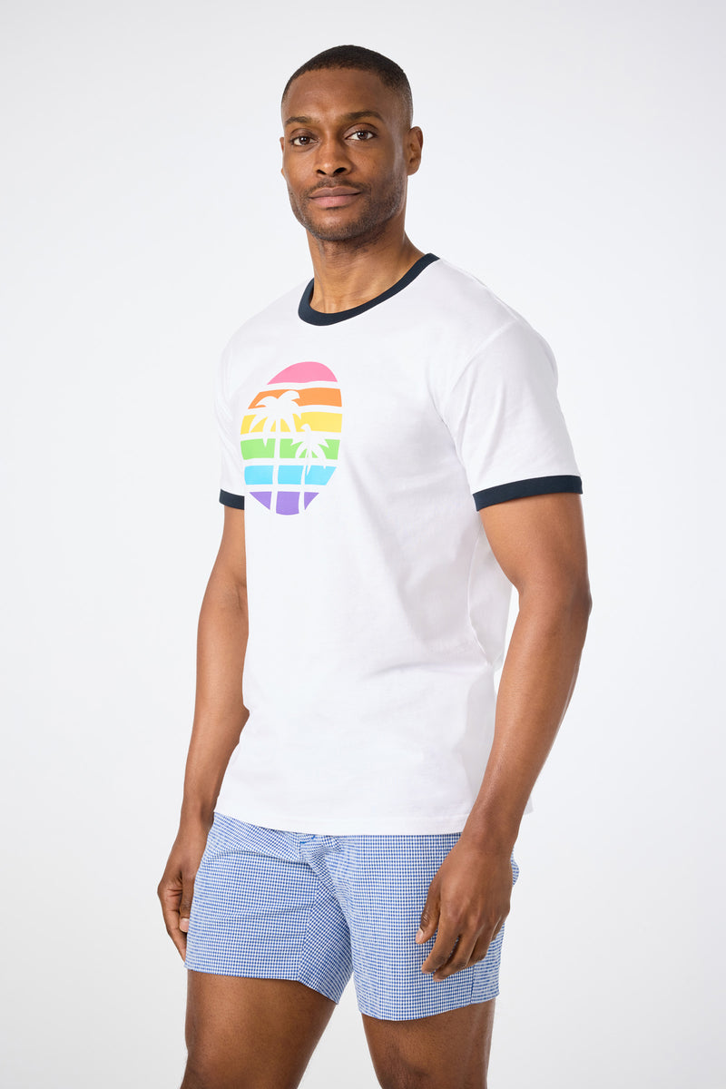 A man stands against a plain background wearing the Short-Sleeve Crewneck T-Shirt - Pride, featuring a rainbow palm tree graphic, and blue checkered shorts. He faces the camera with a neutral expression.