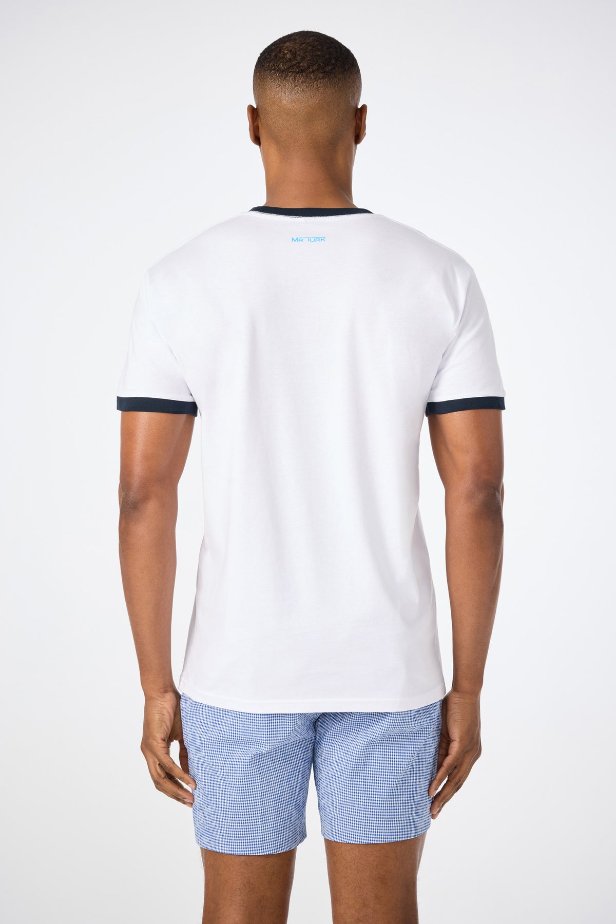 A man faces away from the camera, wearing the Short-Sleeve Crewneck T-Shirt - Pride in white with black trim and light blue checkered shorts, set against a plain white background.