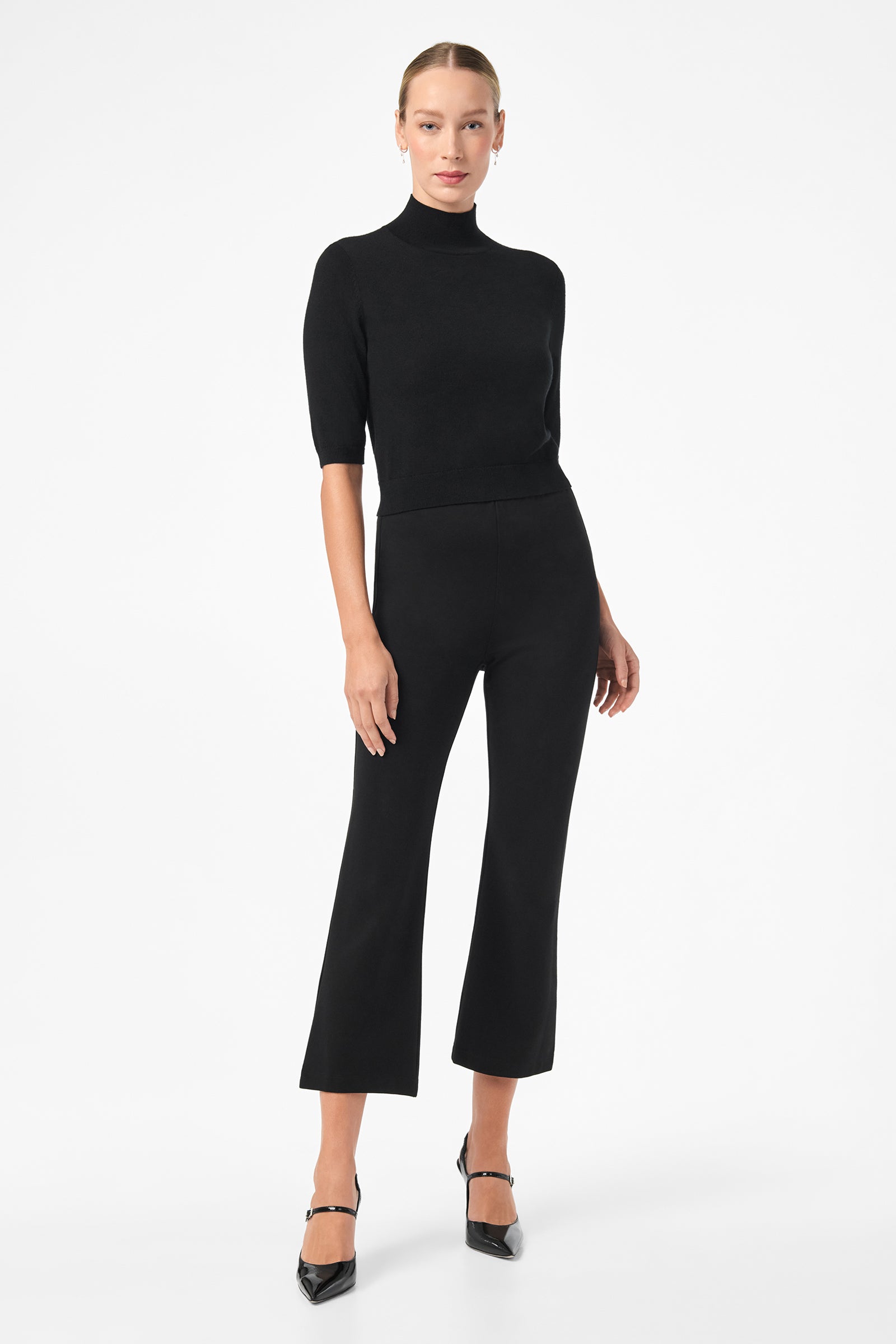 A woman stands against a white background wearing black Sorrel Kick-Flare Pants, a fitted black turtleneck top, and black heels. Her hair is pulled back, and she looks at the camera with a neutral expression.