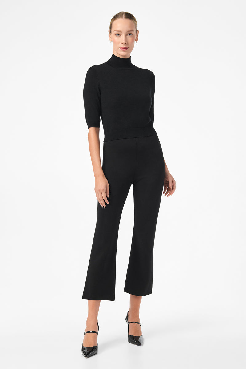 A woman stands against a white background wearing the Mali Mock Neck in black, paired with black cropped pants and heels. She looks forward with her arms relaxed by her sides.