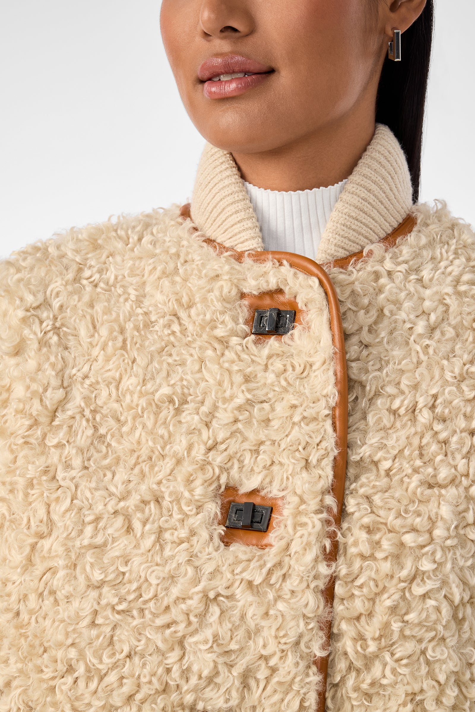 A woman wears the Spartan Coat, a cream-colored curly shearling jacket with large buttons and a ribbed collar, layered over a white turtleneck. Only her lower face and upper torso are shown.