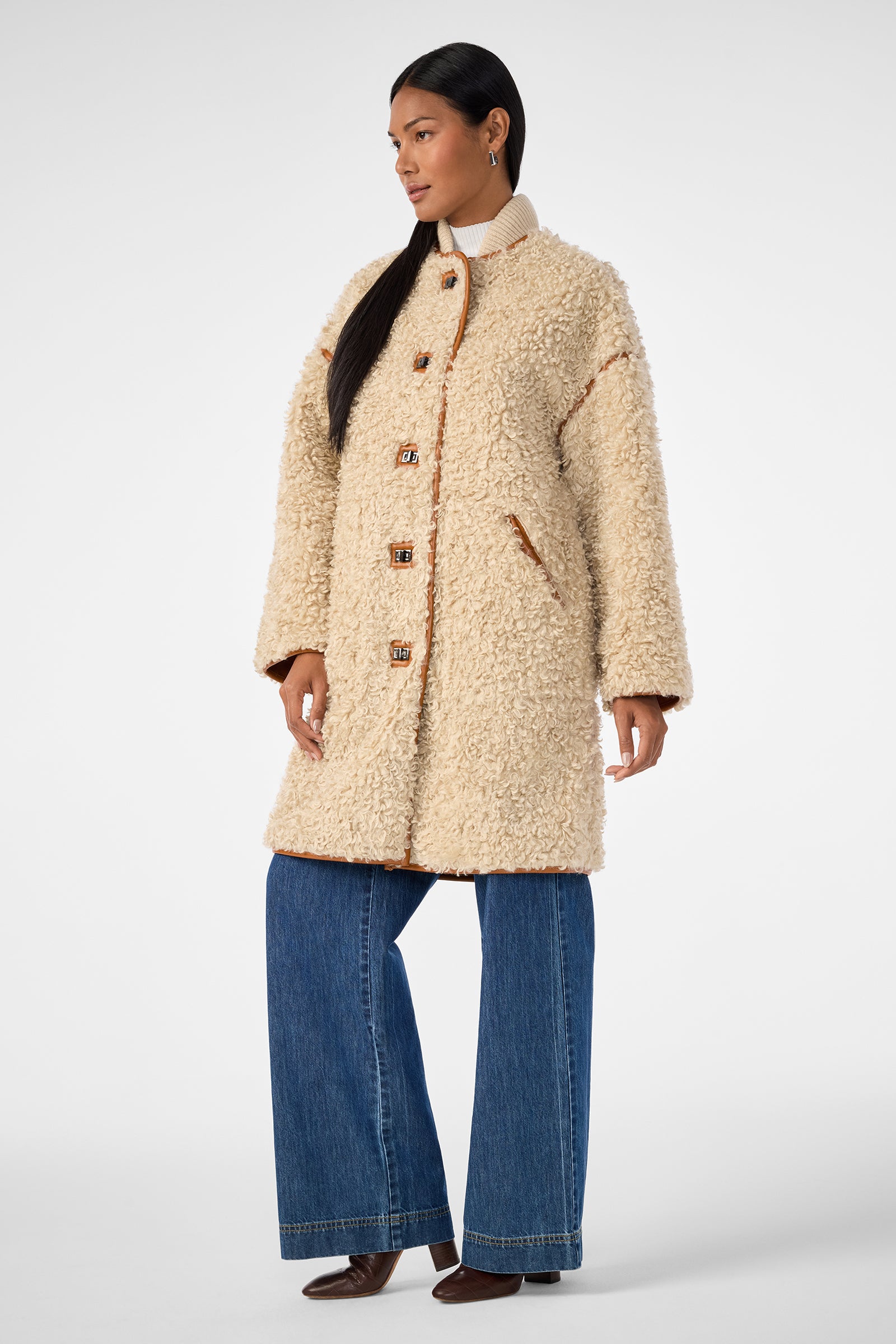 A woman wears the Spartan Coat, a cream-colored, textured button-up shearling layer, over a turtleneck sweater with wide-leg blue jeans and dark brown shoes, standing against a plain white background.
