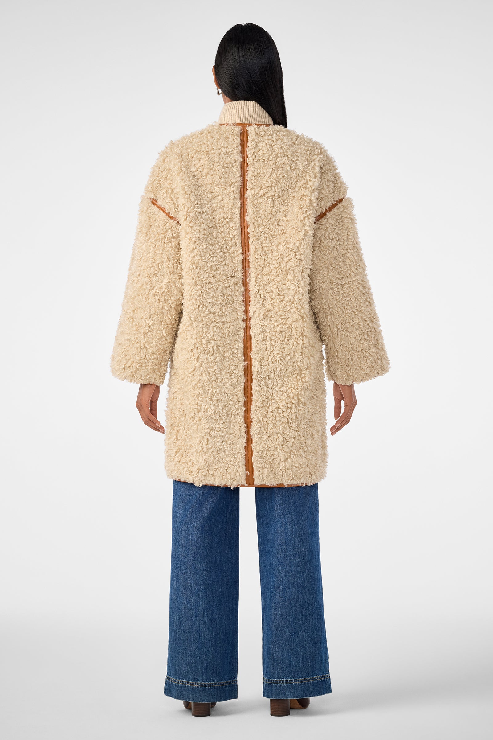 A person with long dark hair, seen from behind, wears the Spartan Coat—a cream-colored, textured shearling coat with tan trim—paired with wide-leg blue jeans and brown shoes against a plain light background.