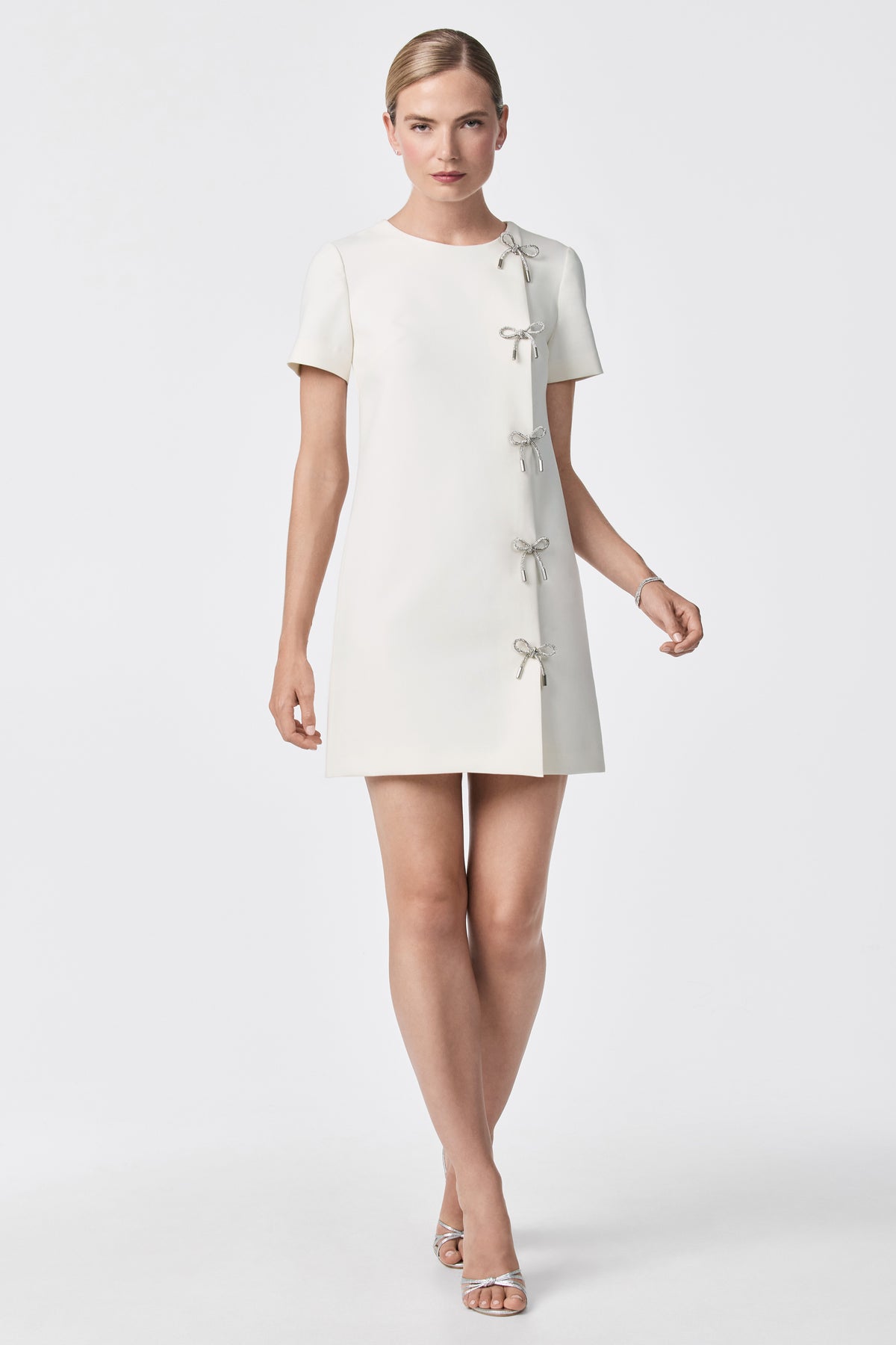 A woman stands against a plain white background wearing the Diamonds Crystal Bows Sheath Dress, a short-sleeved, knee-length white dress with four small bows down the side, paired with silver open-toed sandals. Her hair is pulled back and she has a neutral expression.