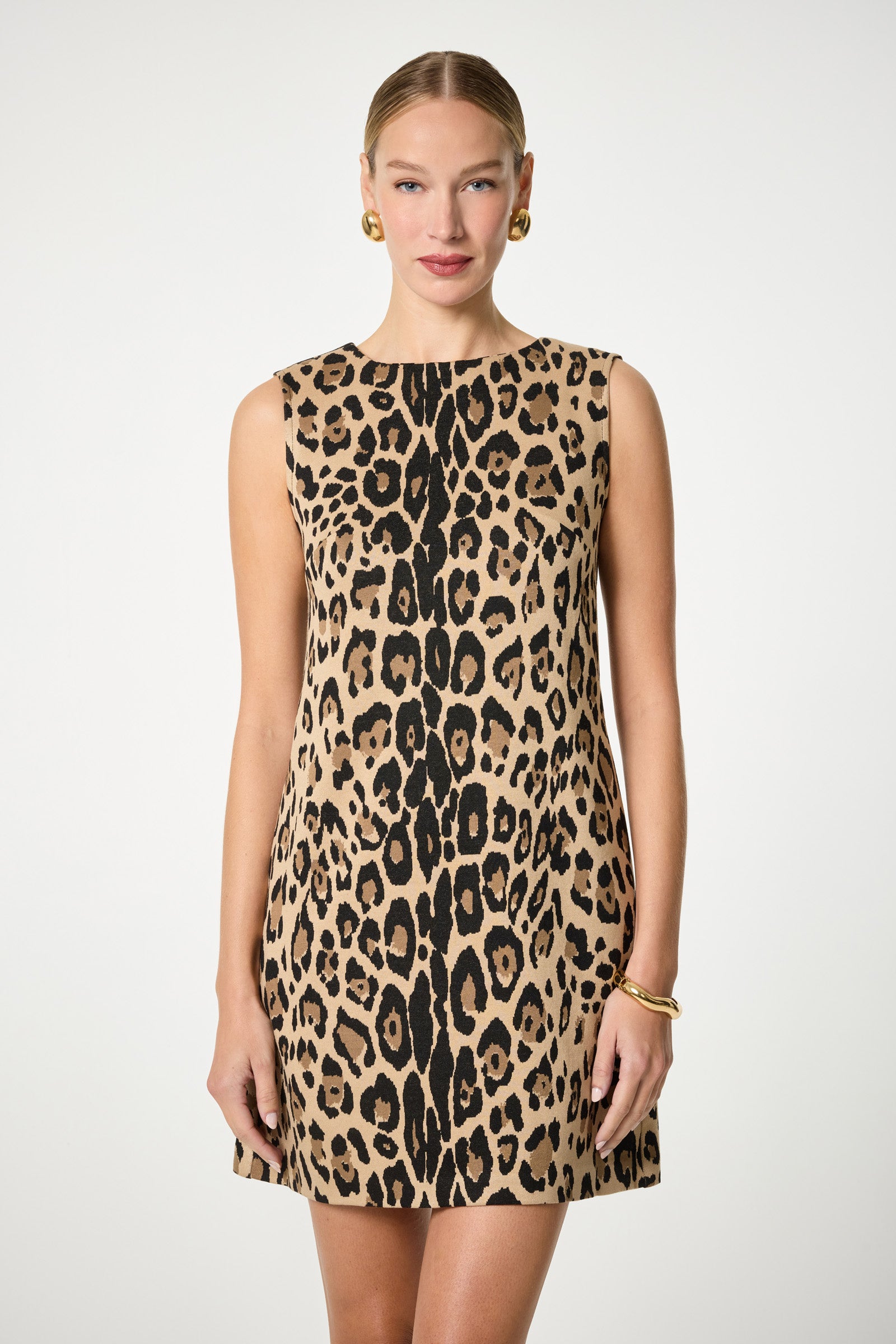 A woman faces forward wearing the Adapted Sleeveless Shift Dress, featuring a bold leopard print and knee-length cut. Her hair is pulled back, and she accessorizes with gold hoop earrings and a bracelet against a plain white background.