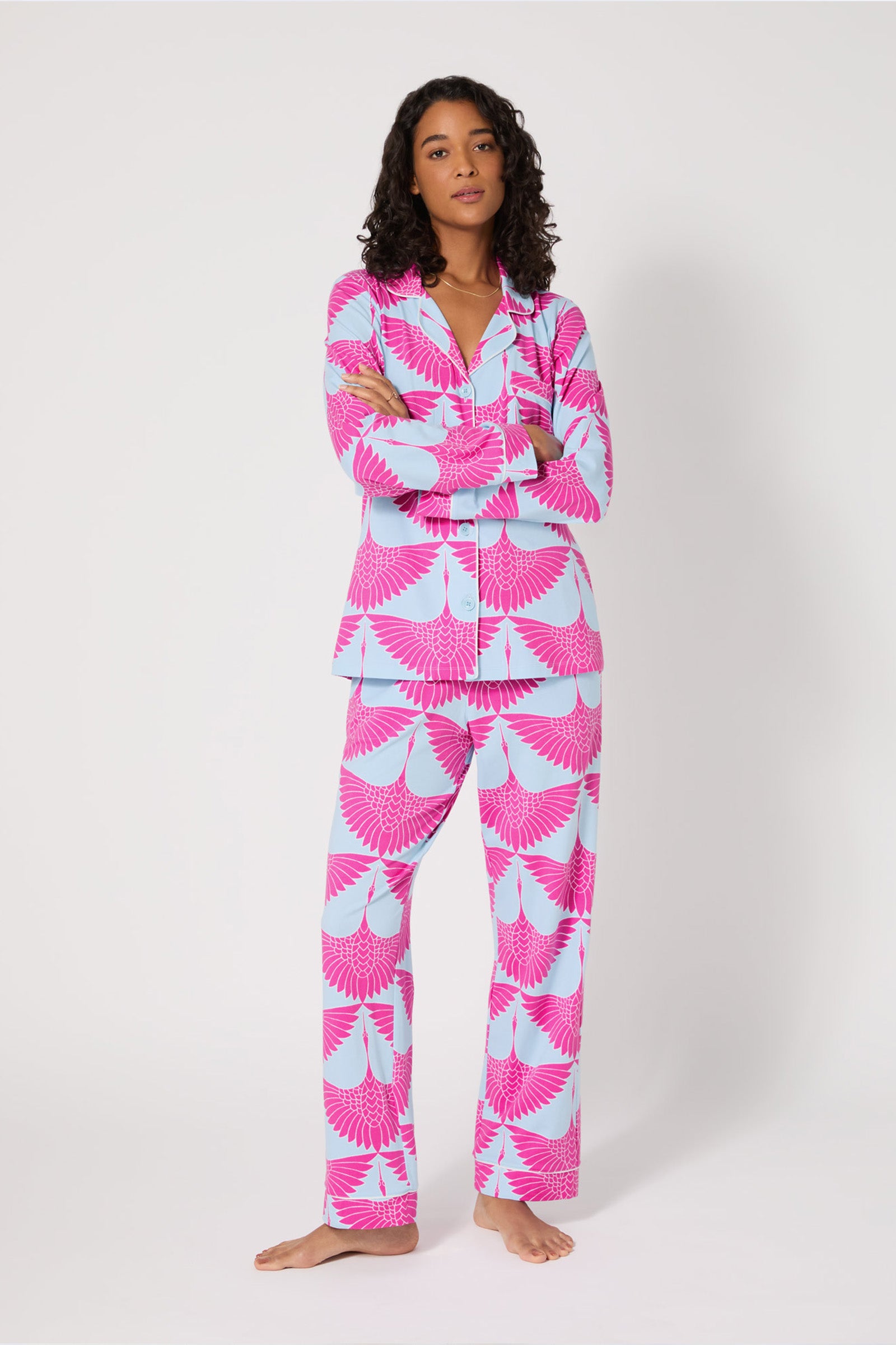 A woman with curly hair stands barefoot against a plain background, wearing the Classic PJ Set - Cranes in MULTI, arms crossed.