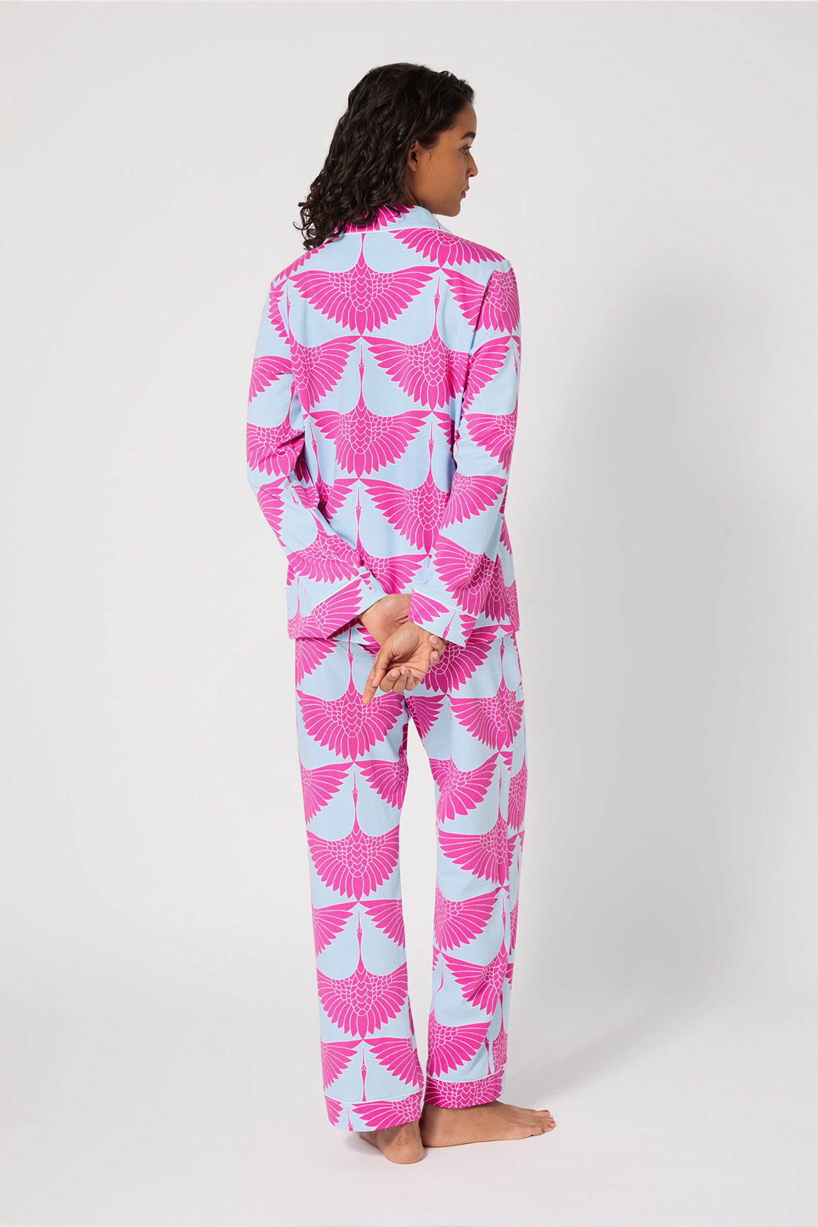 A person stands barefoot, facing away, wearing the Classic PJ Set - Cranes in light blue with bold pink fan patterns. Their hands are clasped behind their back, and they have dark, shoulder-length curly hair.