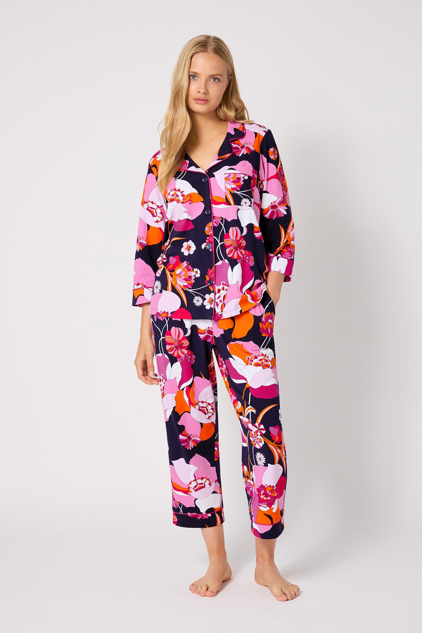 A woman with long blonde hair stands barefoot against a plain background, wearing the Crop Classic PJ Set - Floral Medley, featuring a bold pink, orange, red, and navy blue floral pattern. She appears relaxed.