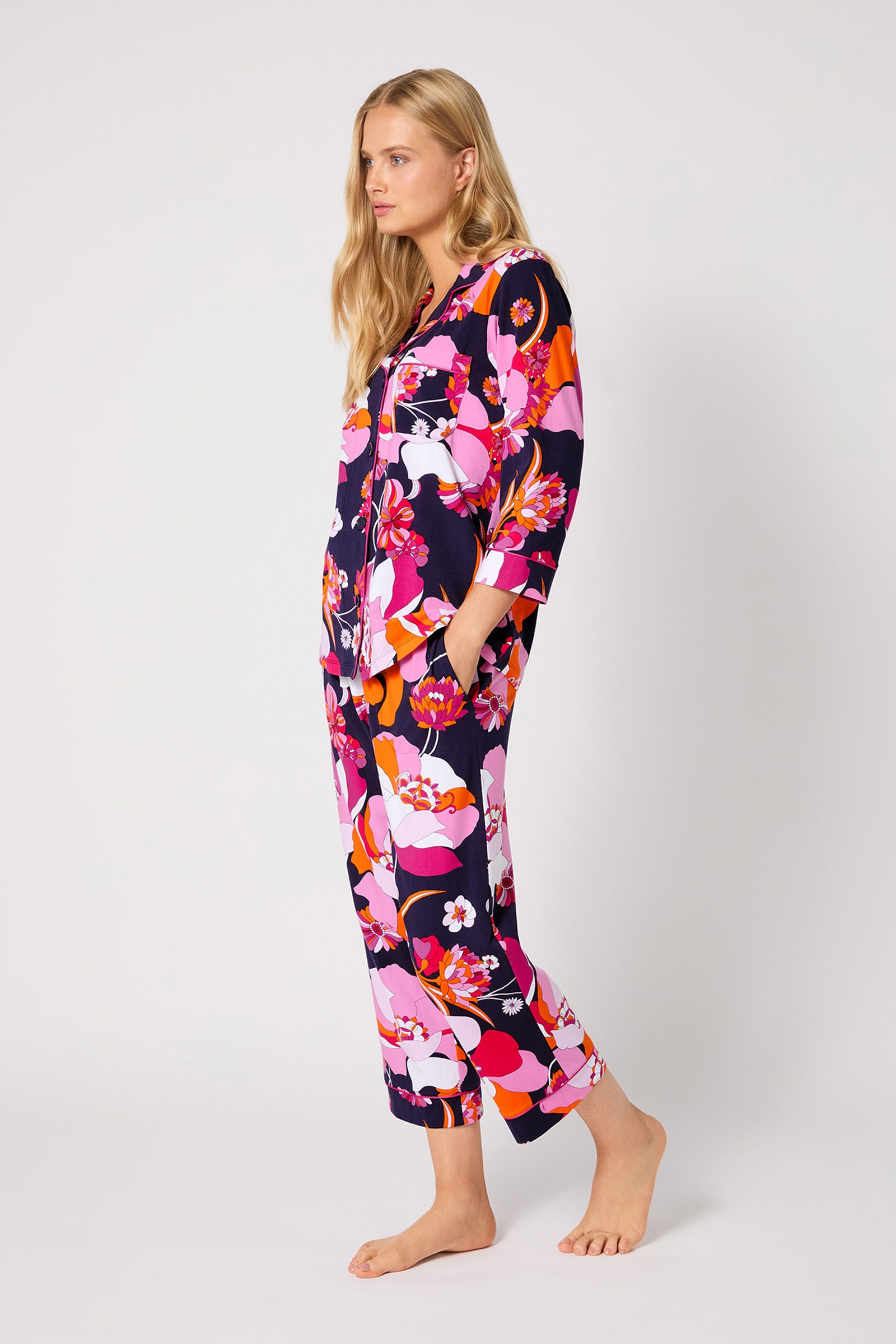 A woman with long blonde hair models the Crop Classic PJ Set - Floral Medley, featuring colorful pink, purple, and orange floral patterns. She stands barefoot against a plain light grey background, looking to the side.