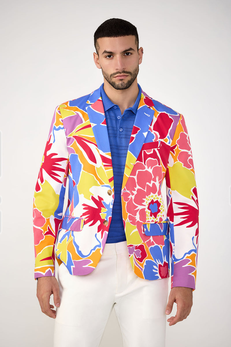 A man wears the Thurston Blazer - Soie Paradis by MR TURK over a blue polo and white pants, standing against a plain light background and looking directly at the camera with a neutral expression.