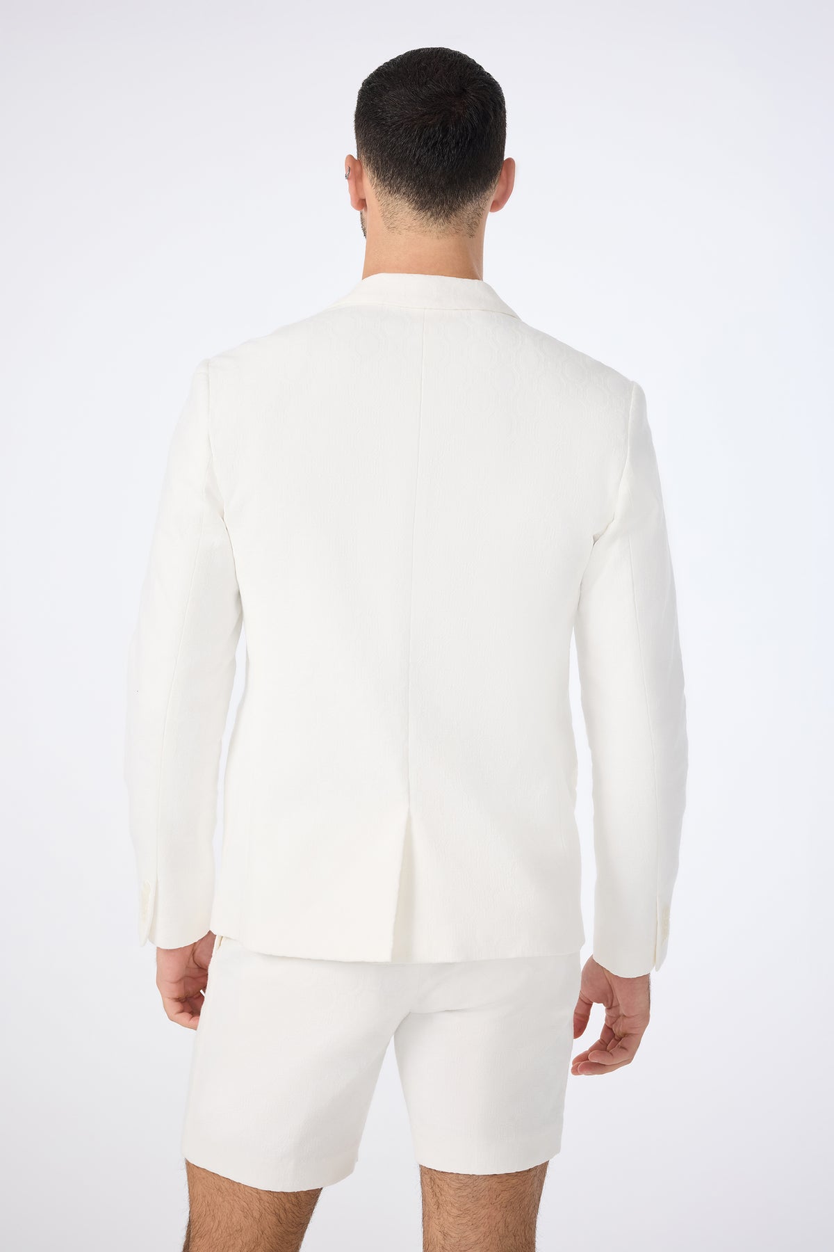 A man faces away from the camera, wearing the Thurston Blazer - Bi-Stretch Cotton and white shorts against a plain light background.