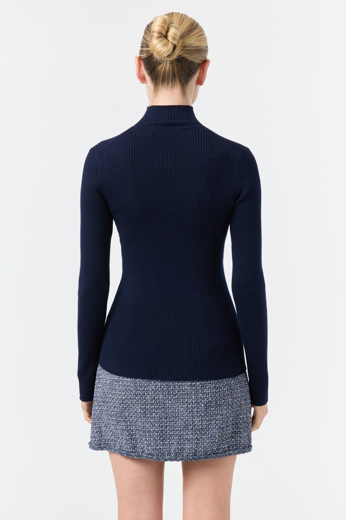 A woman with blond hair in a bun faces away, wearing the Hempstead Turtleneck Sweater in Indigo. The background is plain white.