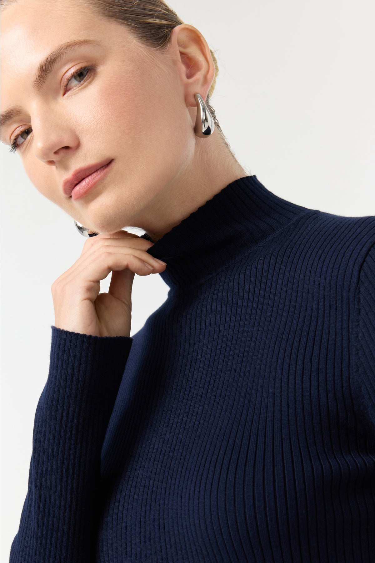 A woman with light skin and blue eyes models the Hempstead Turtleneck Sweater in Indigo, paired with chunky silver earrings, gently touching her neck against a plain light background.