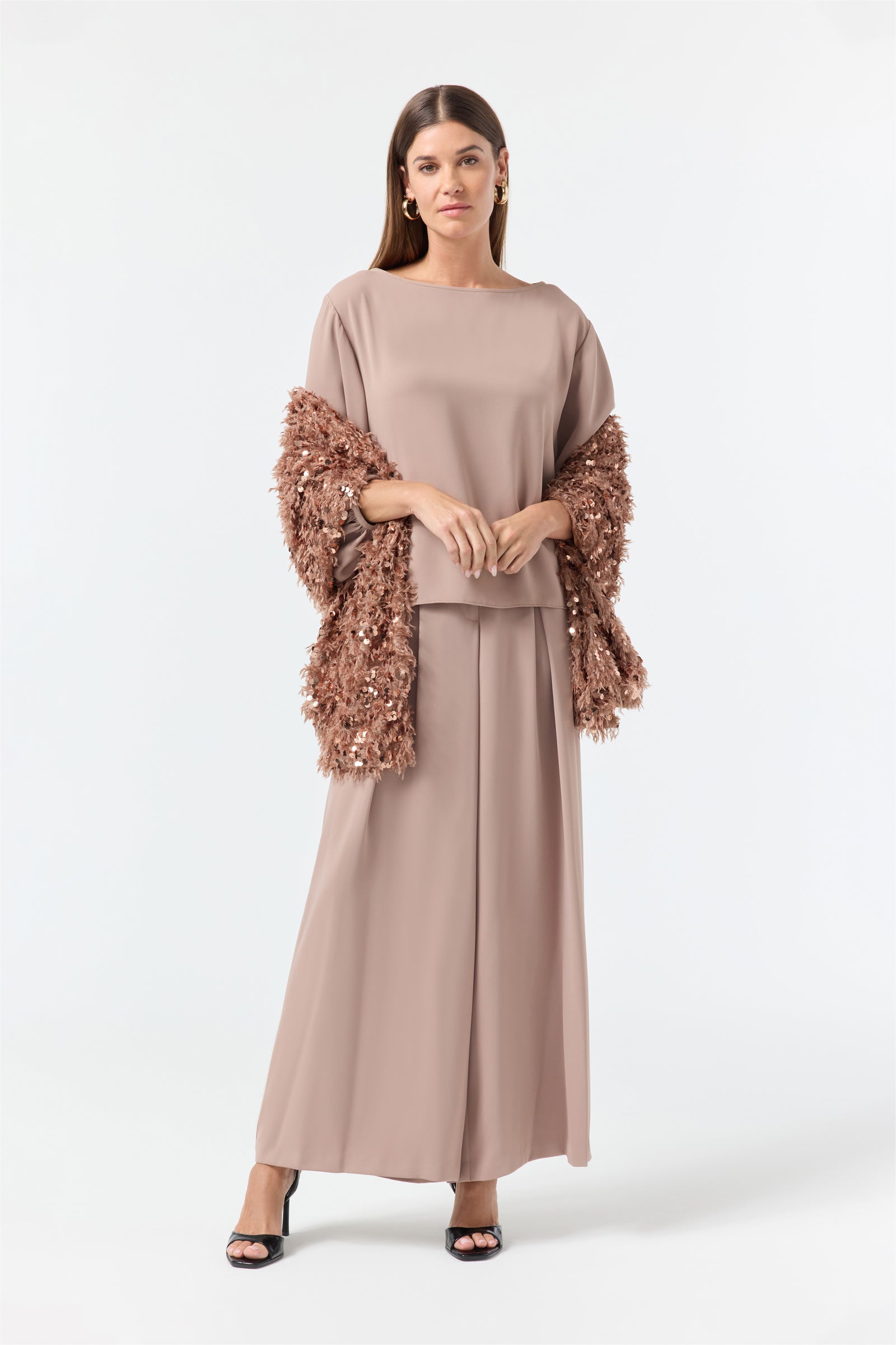 A woman stands on a plain white background wearing a loose taupe outfit with wide-legged pants, a matching top, black heels, and the Fete Faux Fur Stole draped over her arms.