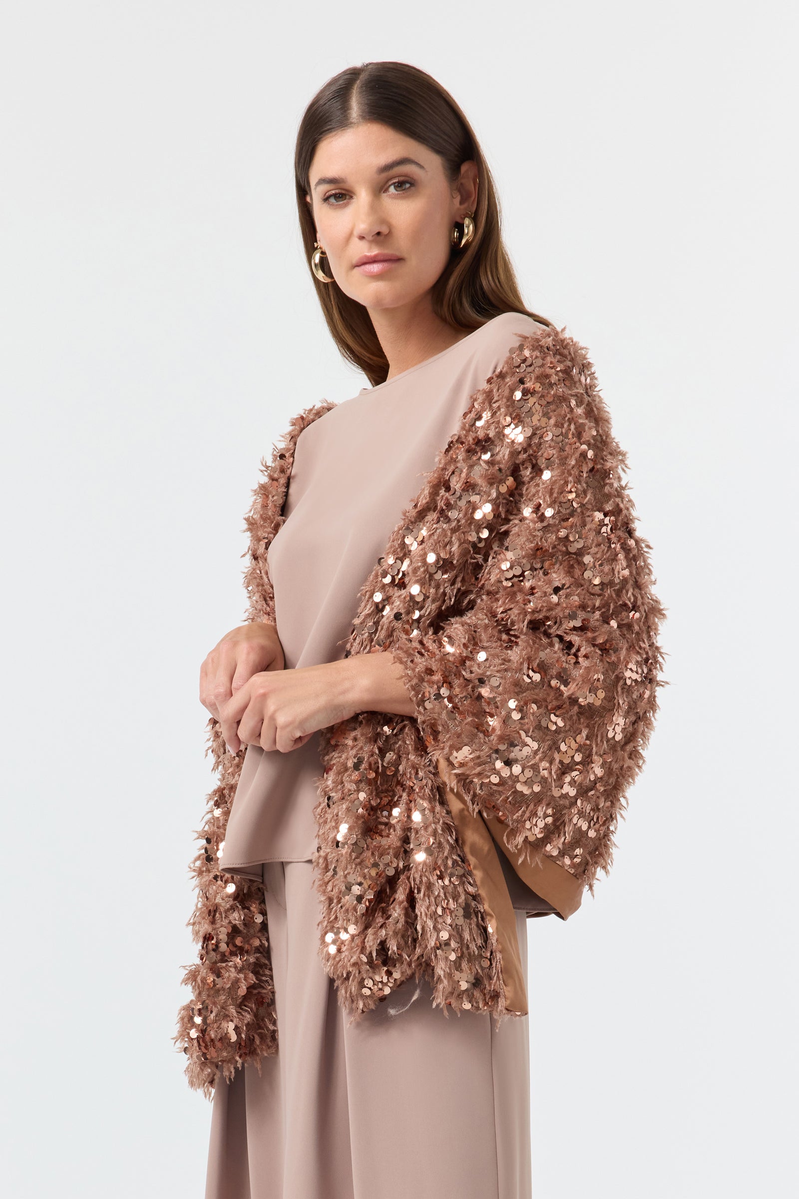 A woman with long brown hair and gold hoop earrings stands facing forward in light taupe clothes, draped in the Fete Faux Fur Stole, a textured, sequined rose-gold shawl. The background is plain white.
