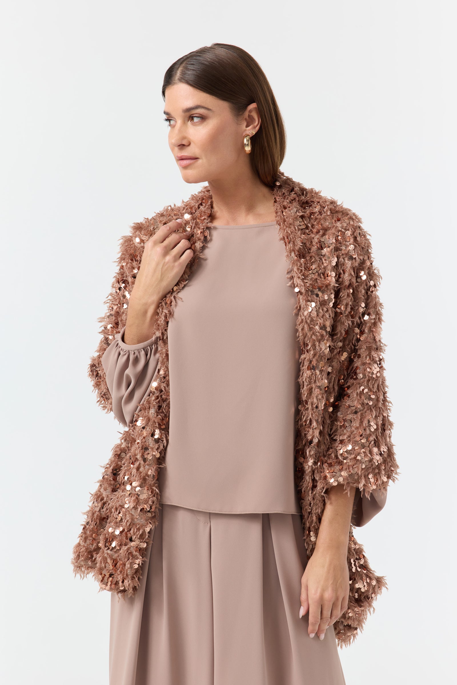 A woman stands against a plain white background, looking to the side, wearing a light brown loose-fit blouse and wide pants set layered with the Fete Faux Fur Stole.