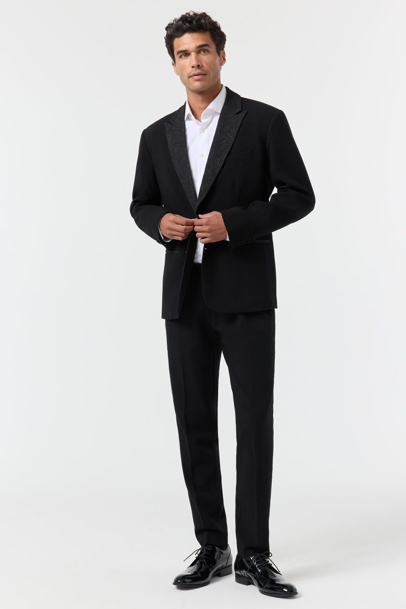 A man stands against a white background, wearing the Miles Blazer over a white dress shirt and black dress shoes. He is buttoning the jacket and gazes slightly upward with a neutral expression.