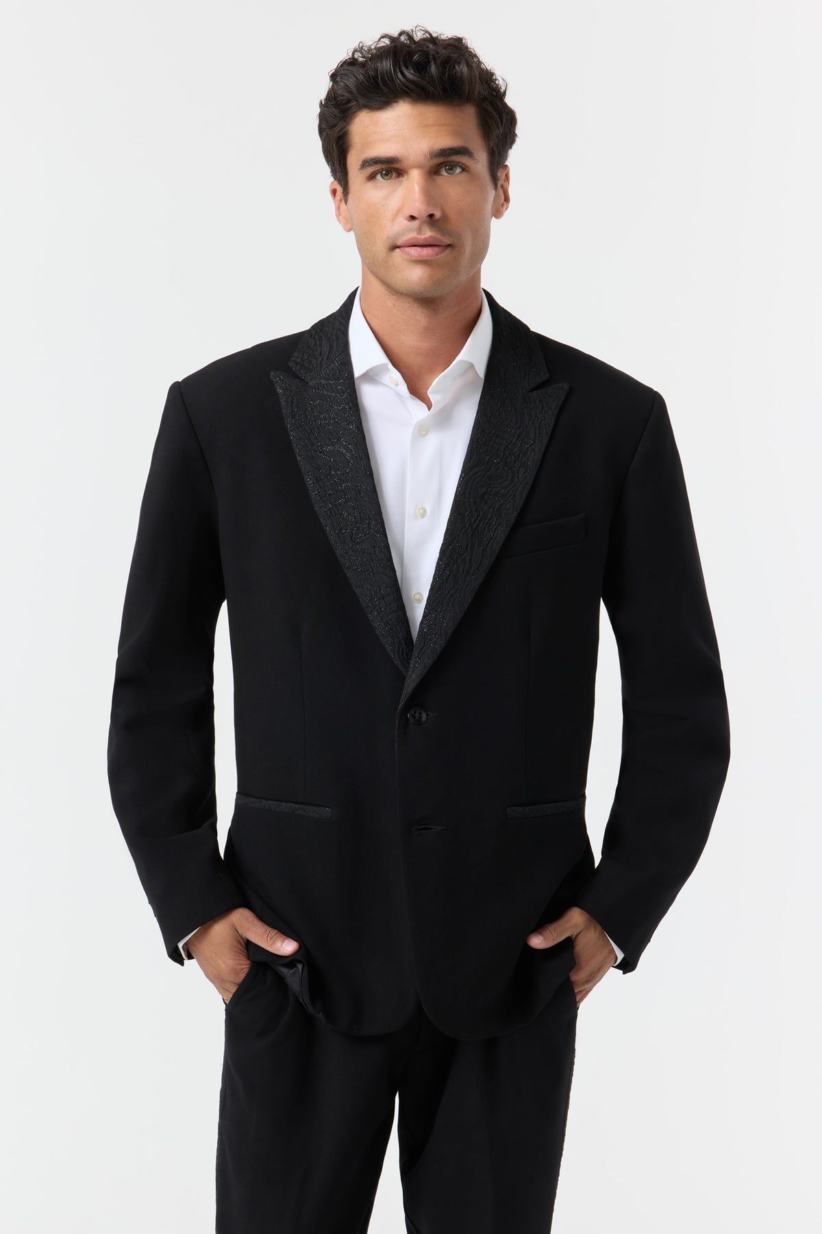 A man with dark hair models the Miles Blazer—black suit jacket and white dress shirt—standing forward with hands in his pockets against a plain white background.