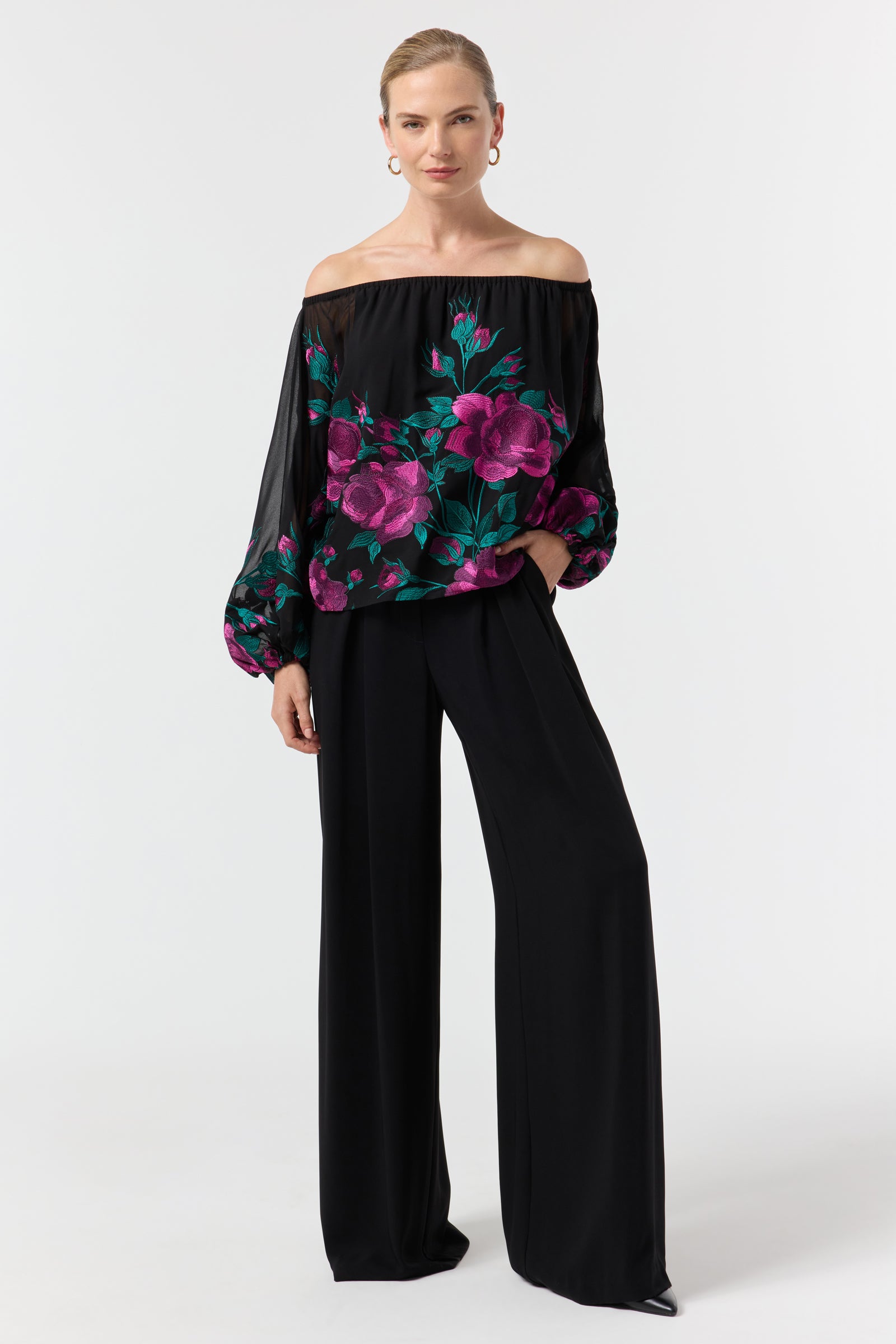 A woman models the Soho 2 Embroidered Top—an off-the-shoulder black blouse with vibrant pink and green floral embroidery—paired with wide-leg black pants, hoop earrings, and her hair pulled back, standing against a white background.