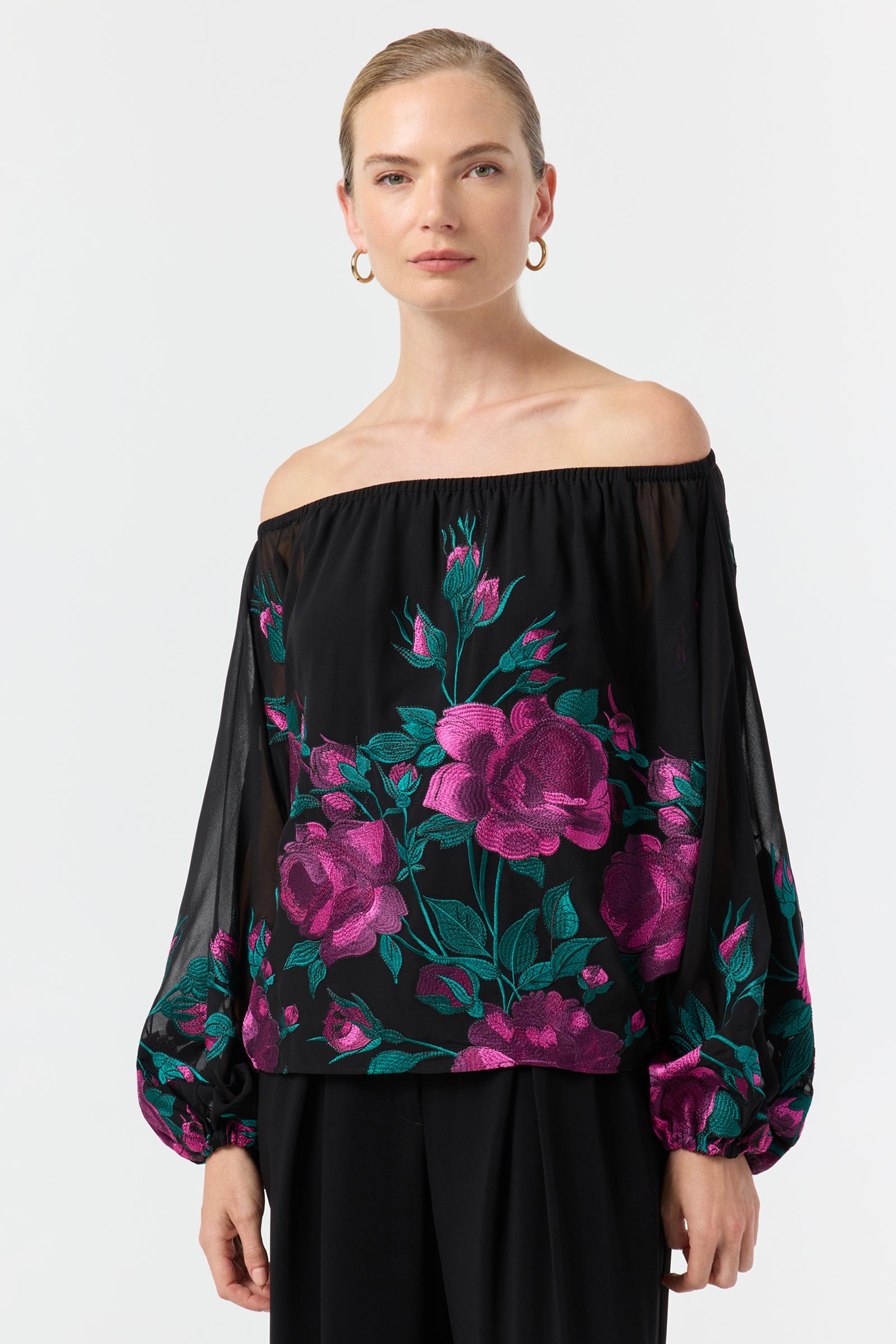 A woman wears the Soho 2 Embroidered Top—an off-the-shoulder black blouse with sheer sleeves and vibrant pink and green floral embroidery—paired with black pants, standing against a plain white background.