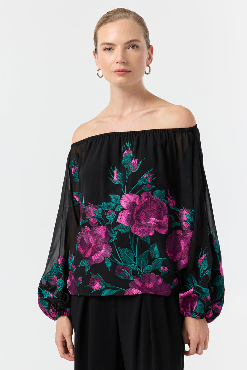 A woman wears the Soho 2 Embroidered Top—an off-the-shoulder black blouse with sheer sleeves and vibrant pink and green floral embroidery—paired with black pants, standing against a plain white background.