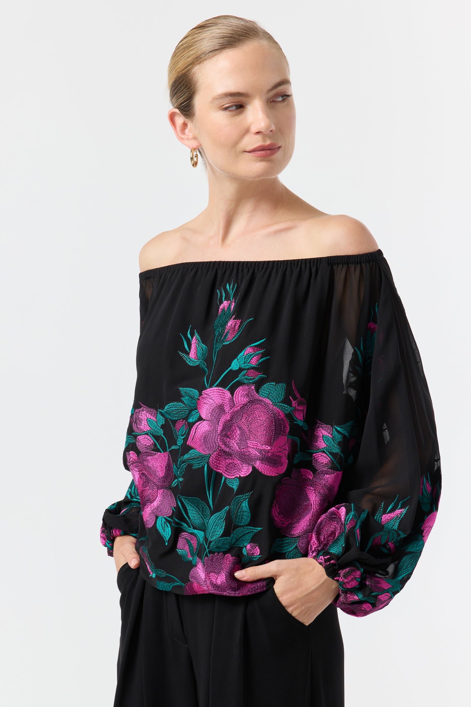 Woman wearing the Soho 2 Embroidered Top—an off-the-shoulder black blouse with pink and green floral embroidery and long sleeves—paired with black pants. Her light hair is pulled back as she smiles softly, looking to the side.