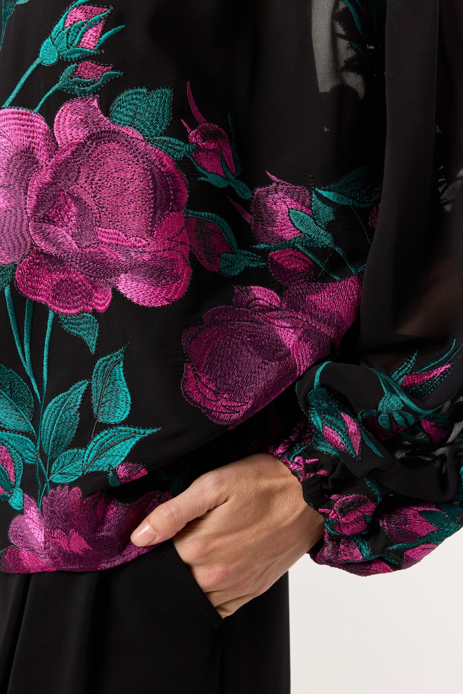 A close-up of a person wearing the Soho 2 Embroidered Top, a sheer black blouse adorned with bright pink embroidered roses and green leaves, with their hand resting on their hip.